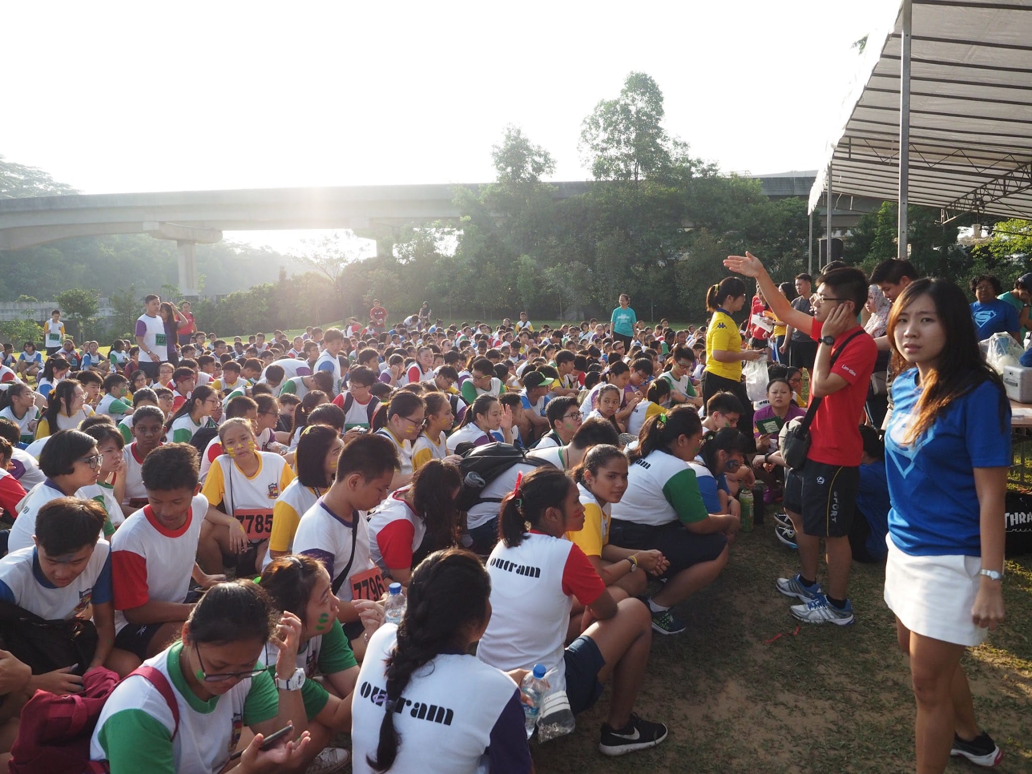 Large crowd of participants in colorful race bibs sitting on grass outdoors near a bridge at a running event.