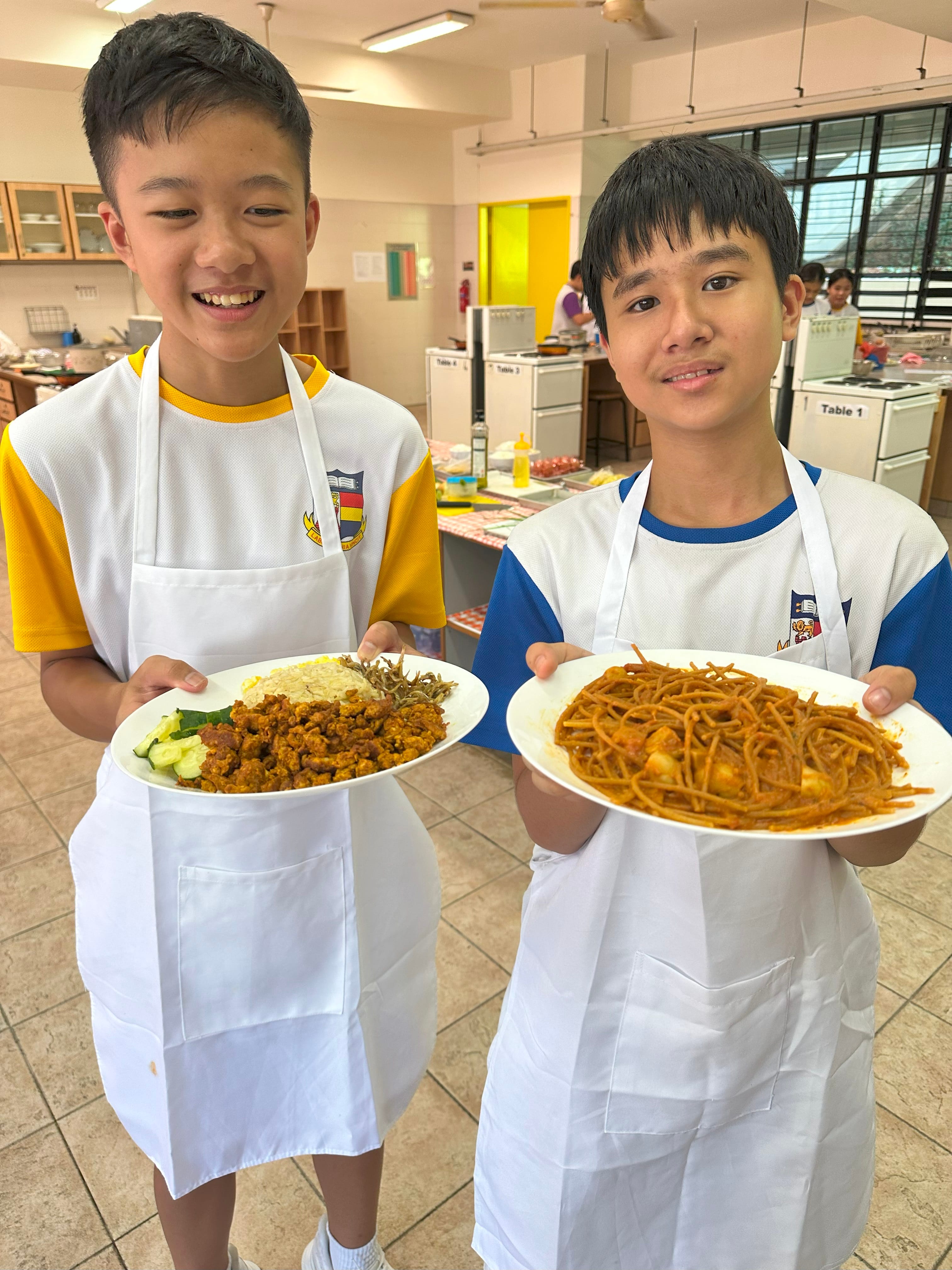 Two smiling boys in white aprons hold plates of cooked rice with meat and noodles in a school kitchen classroom.