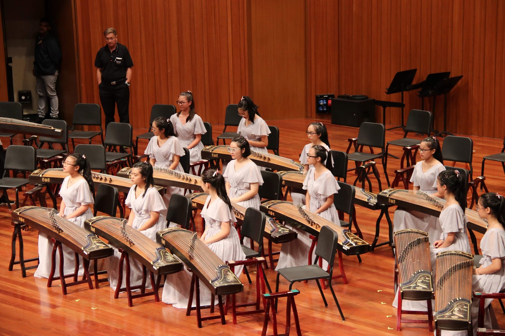 Young women in light gray dresses stand beside guzheng instruments on a wooden concert stage.