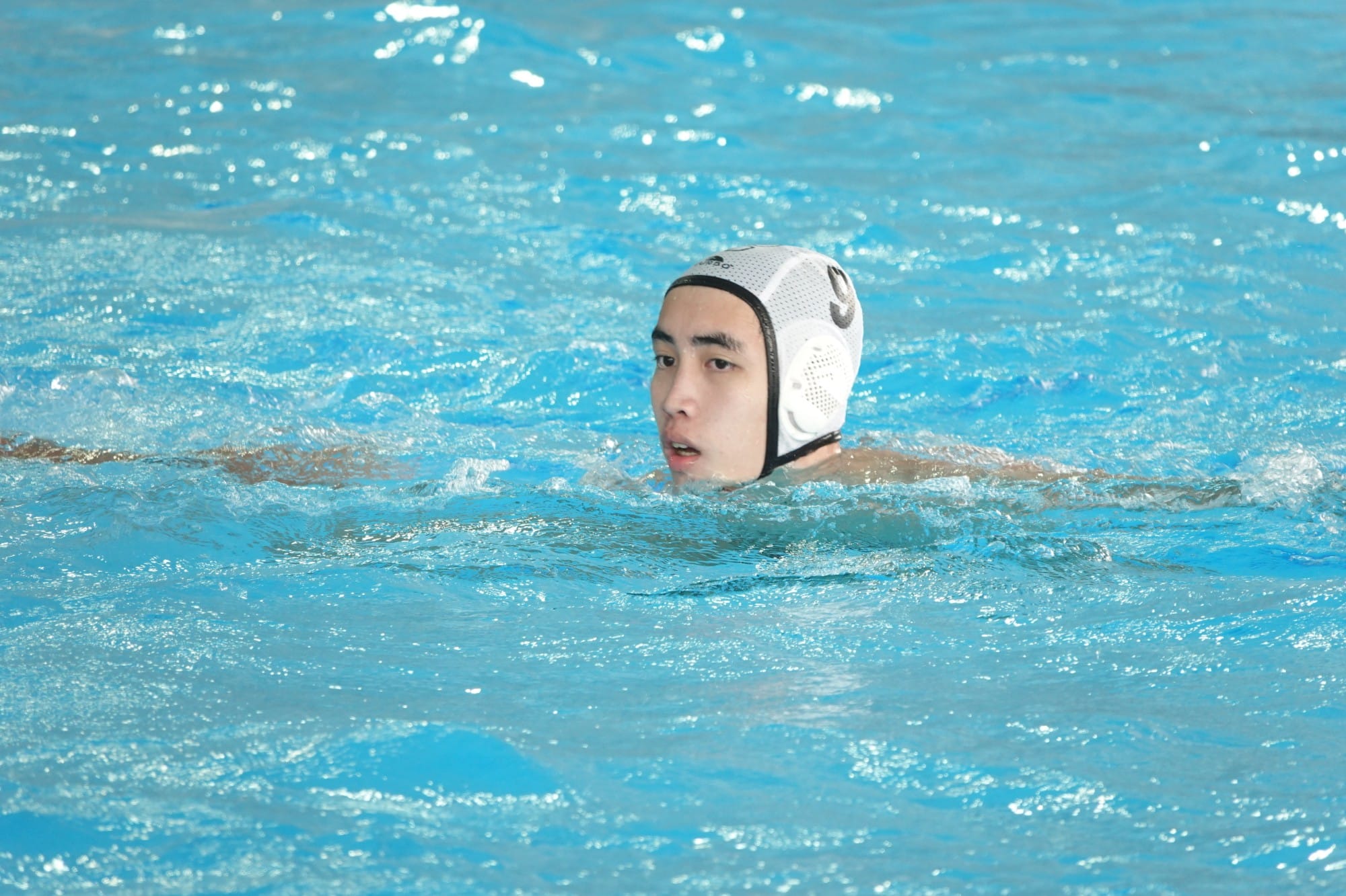 A young man wearing a white water polo cap treads water in a bright blue pool.
