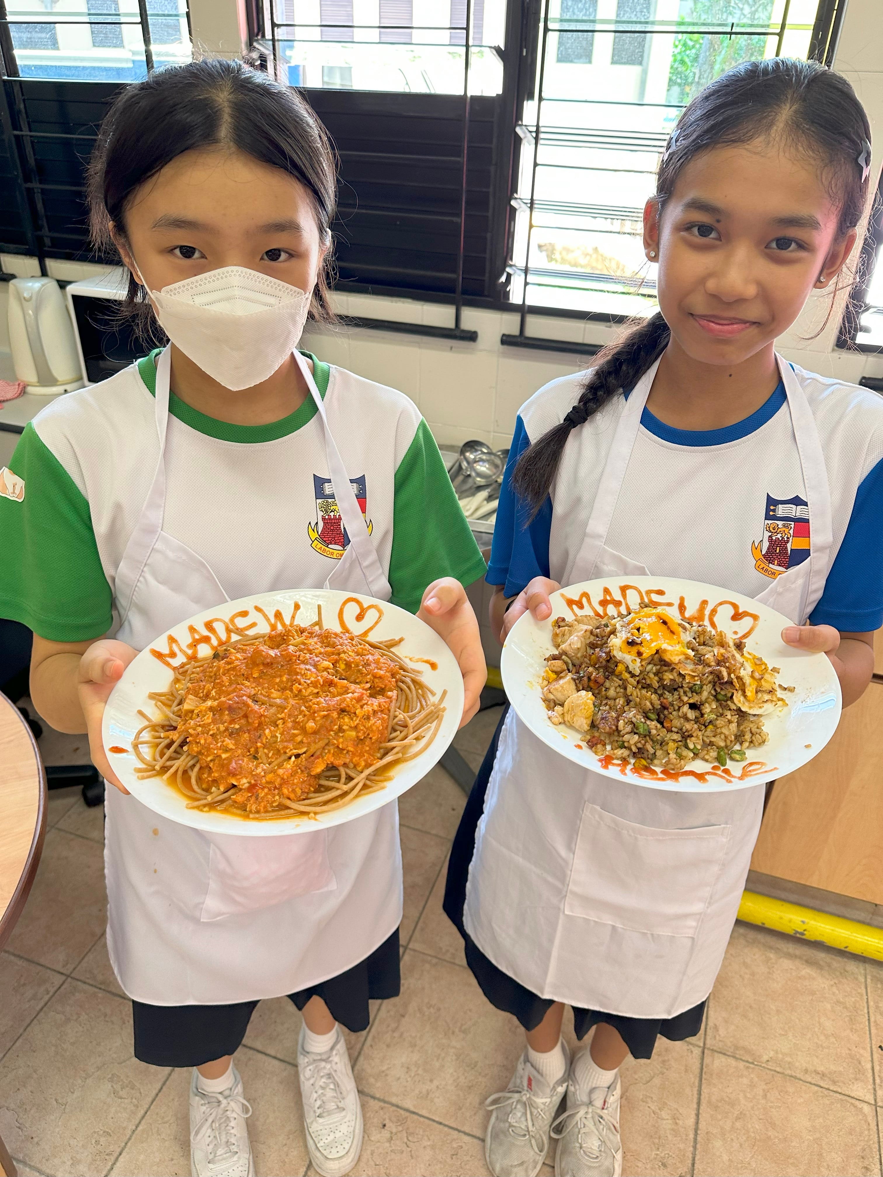 Two young girls in school uniforms and white aprons hold plates of homemade spaghetti and fried rice in a classroom kitchen.