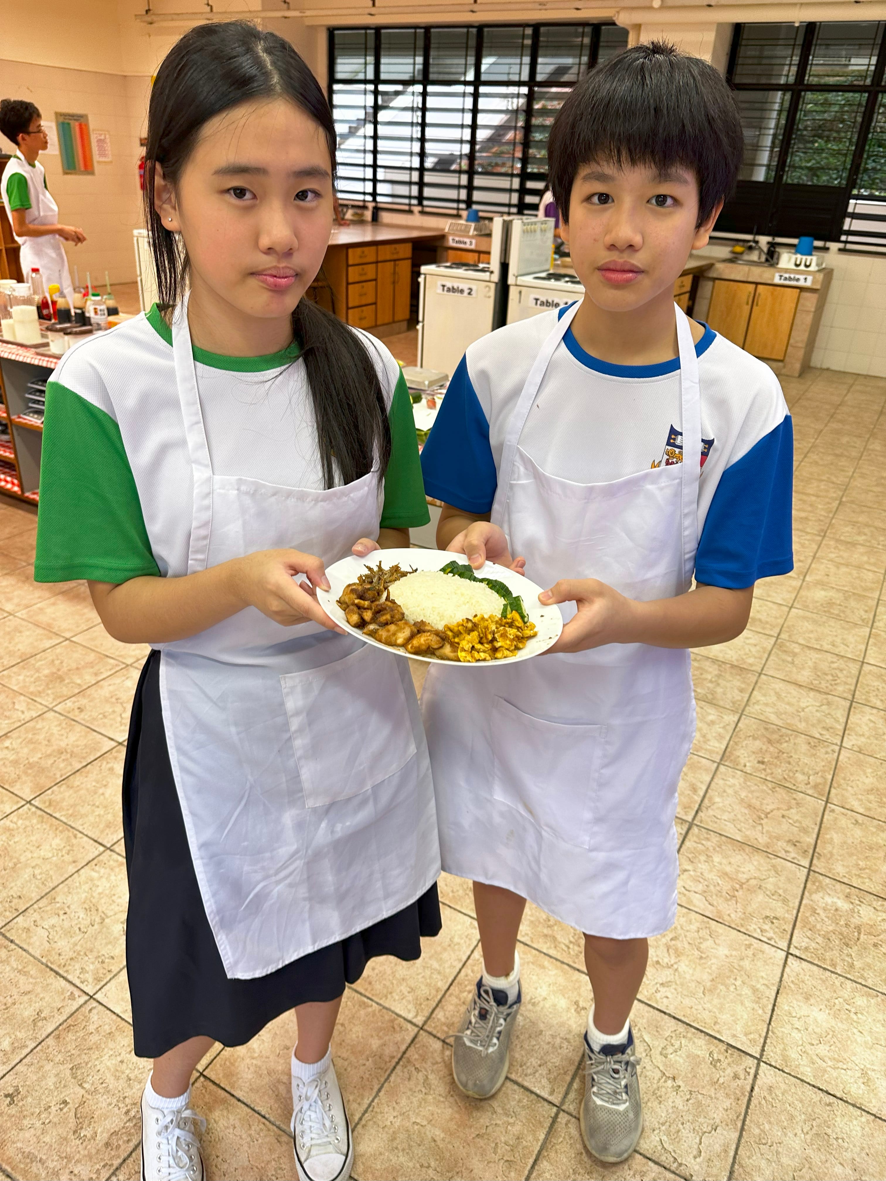 Two school children in white aprons hold a plate of rice with cooked dishes in a classroom kitchen.