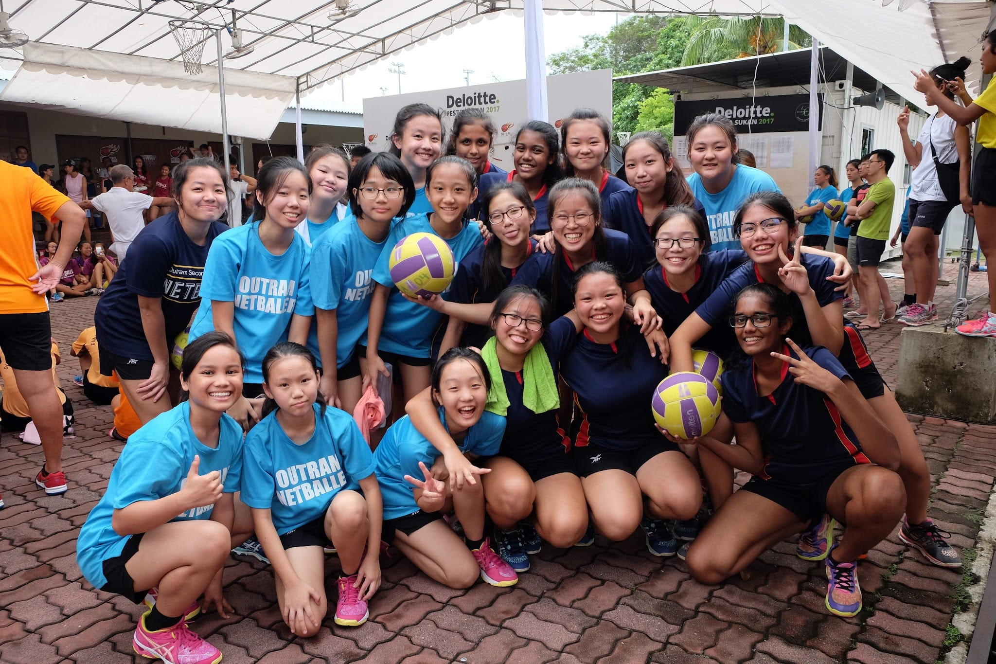 Two girls' netball teams in blue and navy jerseys pose together holding netballs at a 2017 Deloitte event.