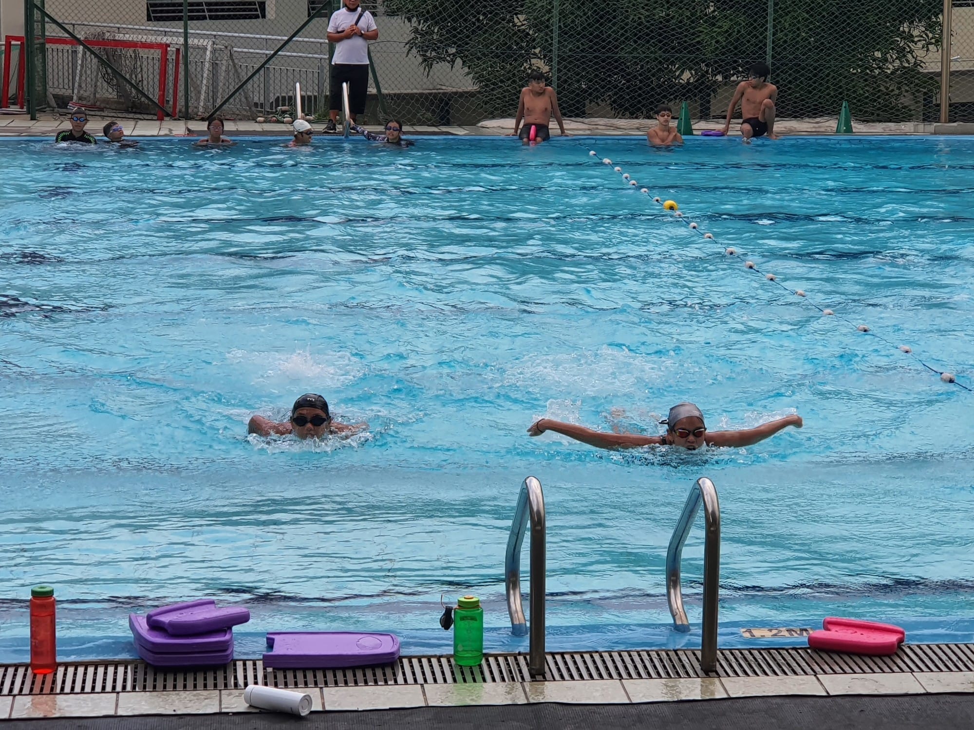 Two children swim butterfly stroke in an outdoor pool while others watch from the edges nearby.