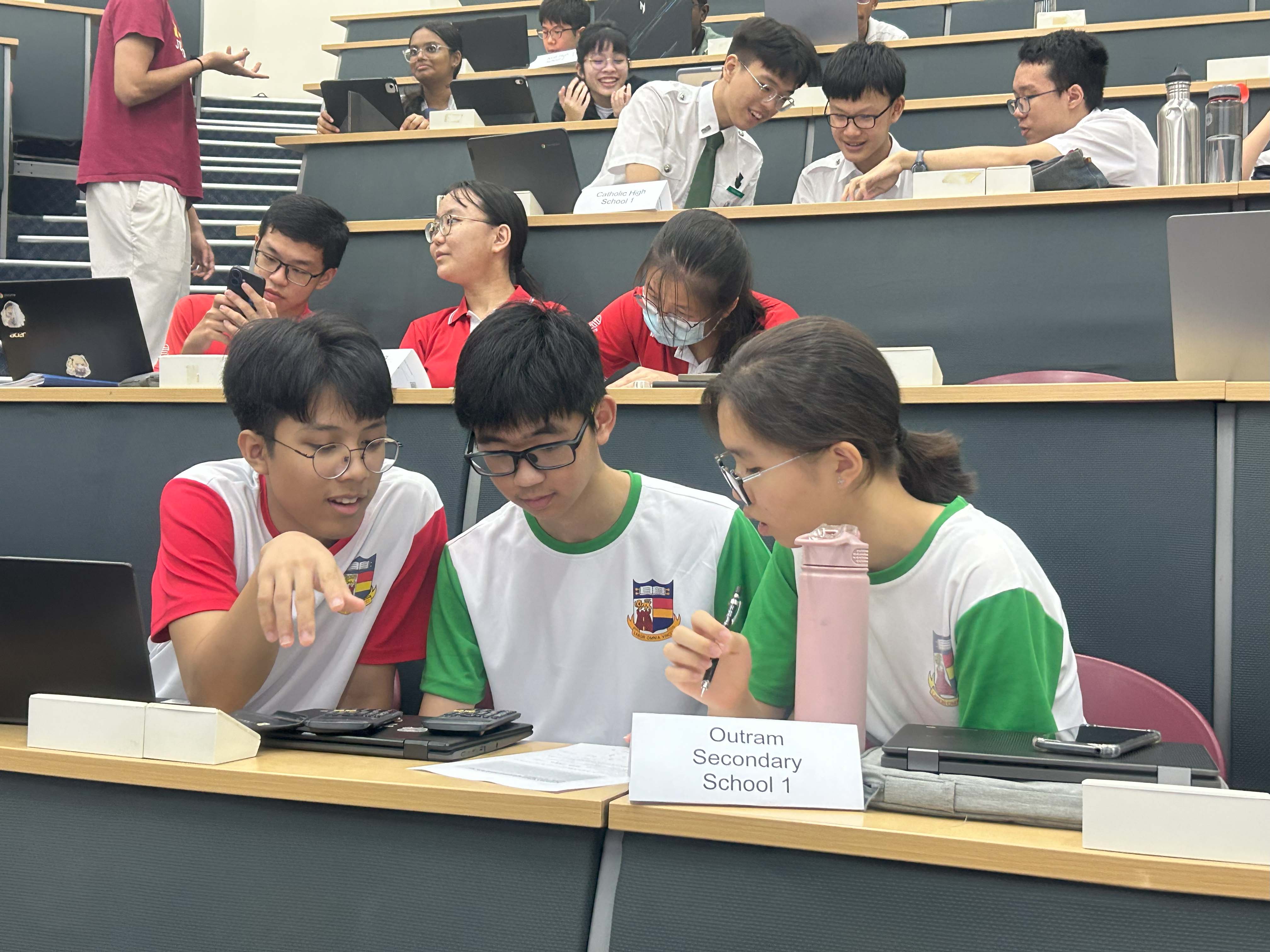 Three students in uniform huddle around a laptop at a desk labeled Outram Secondary School 1 in a lecture hall.