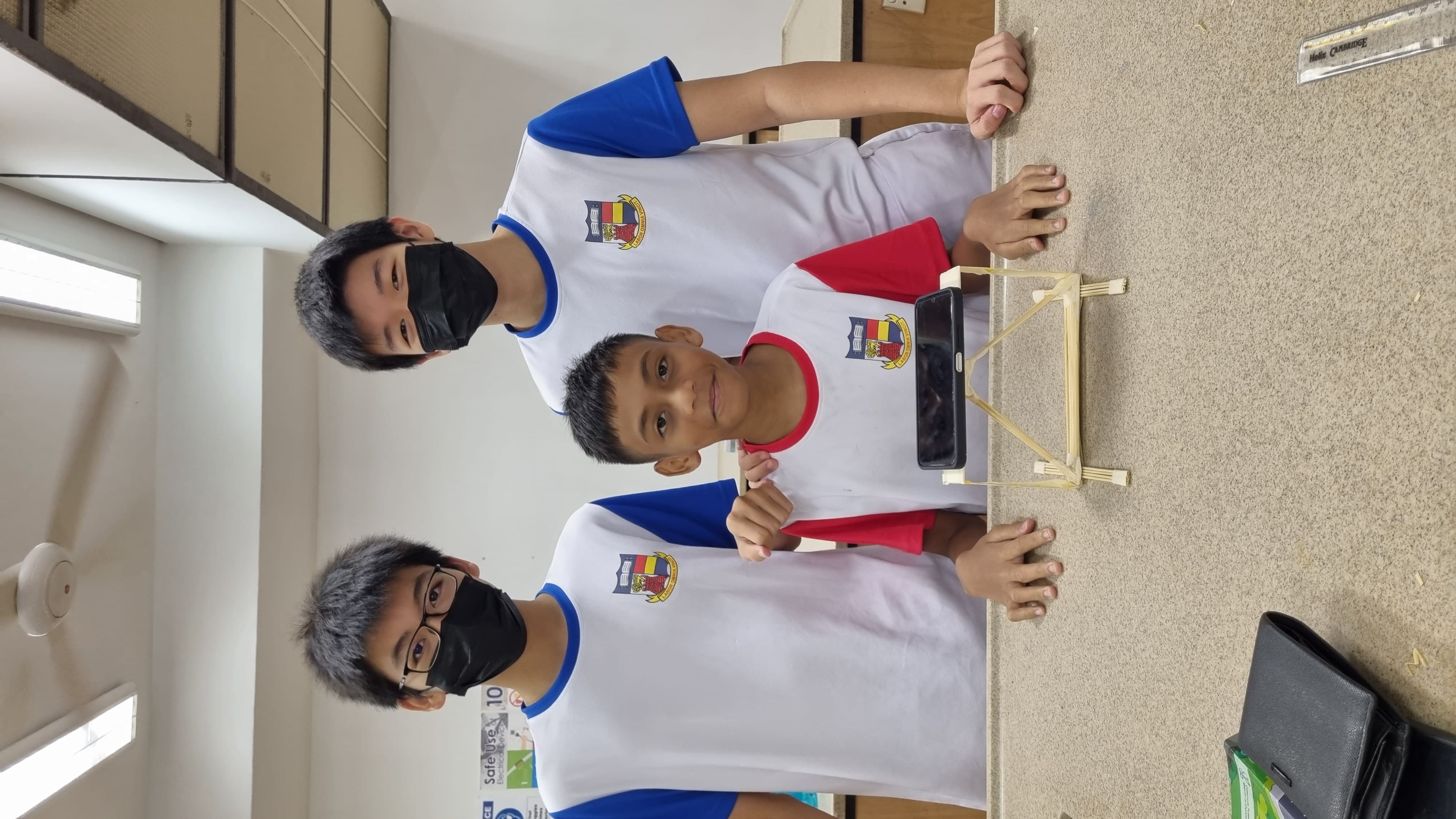 Three school boys in uniforms pose with a small wooden stick bridge model holding a phone on a desk.