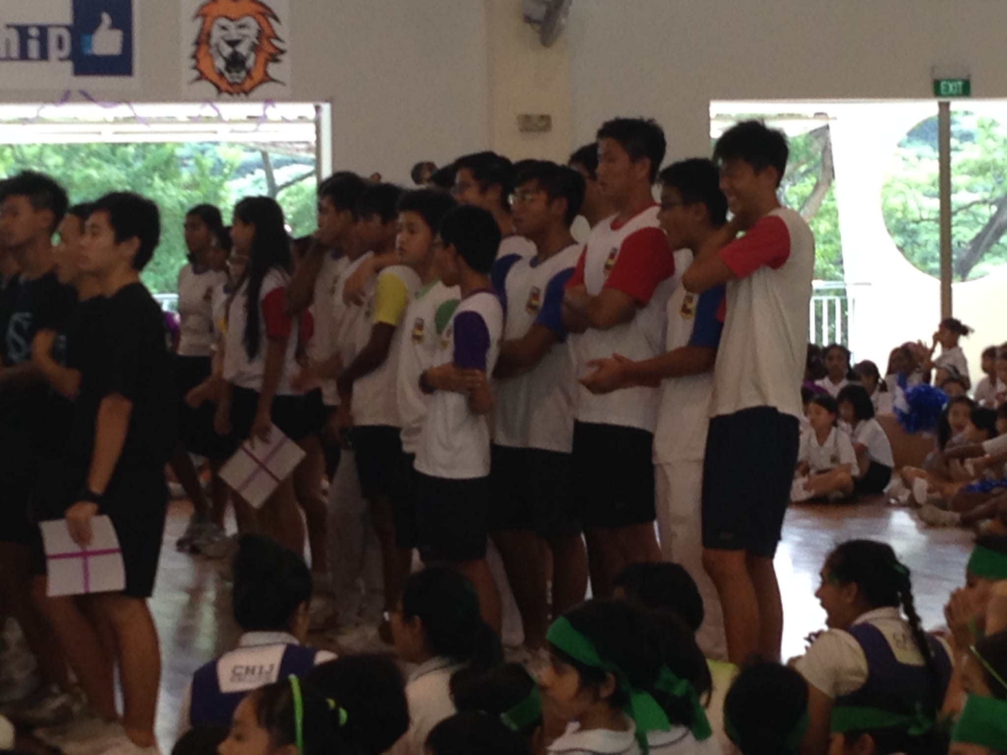 Students in white and colorful shirts stand in a line inside a school hall with seated children watching nearby.