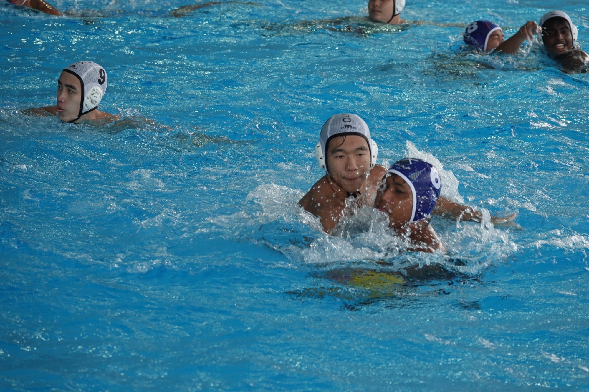 Several water polo players in white and blue caps compete in a bright blue pool during a match.