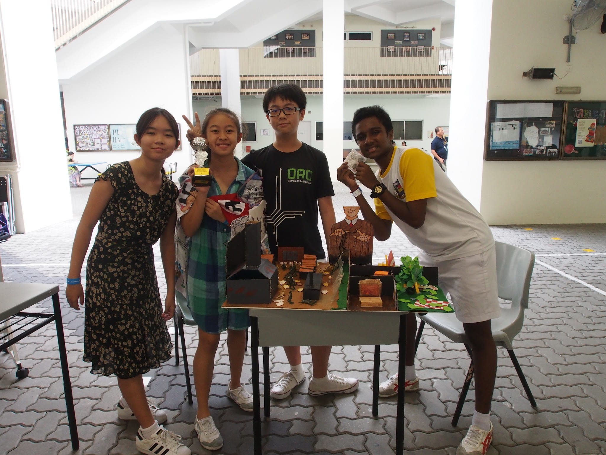 Four students pose proudly beside a diorama project on a table, one holding a small trophy, in a school atrium.