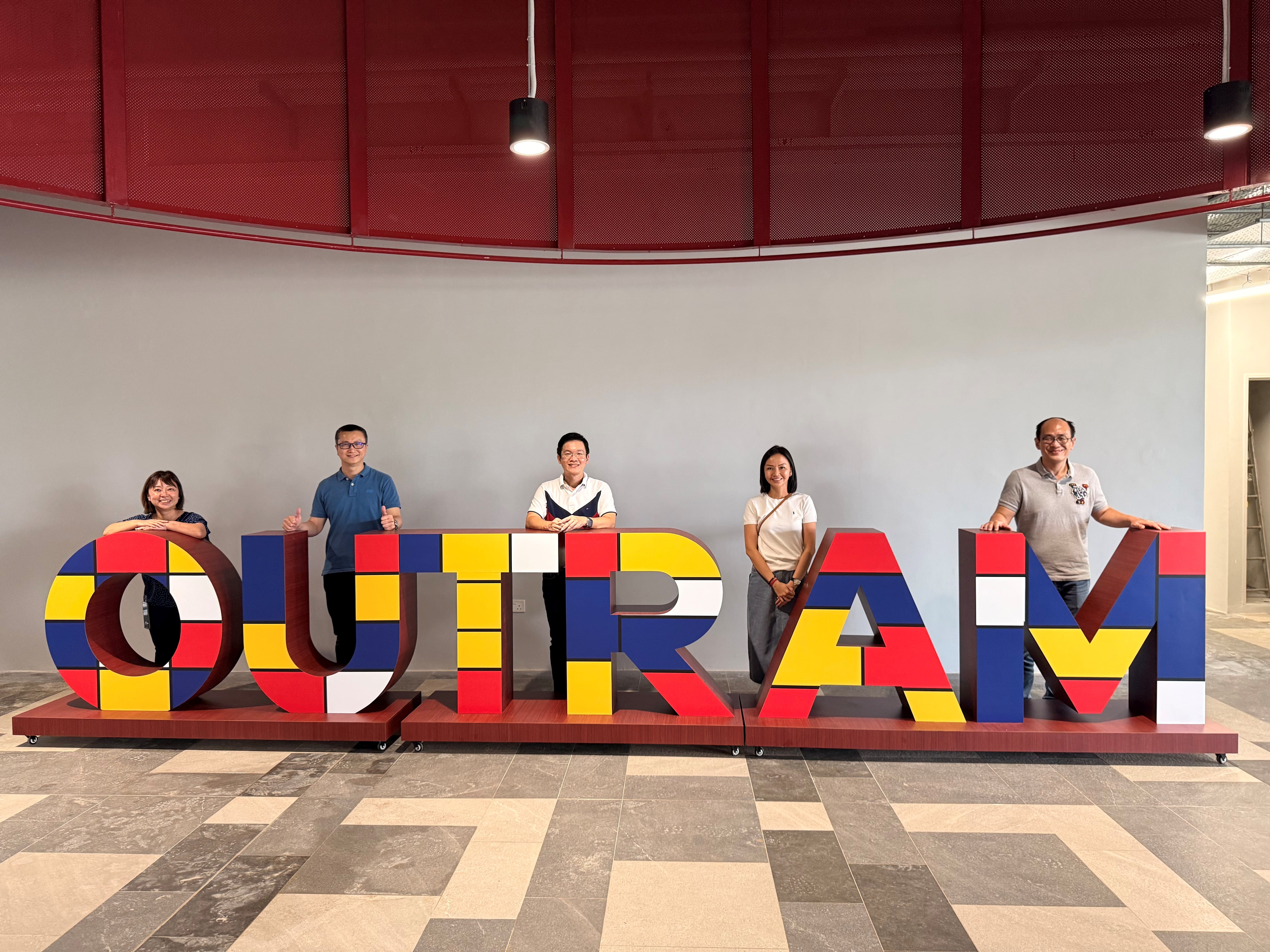 Five people standing behind large colorful block letters spelling OUTRAM in an indoor space.