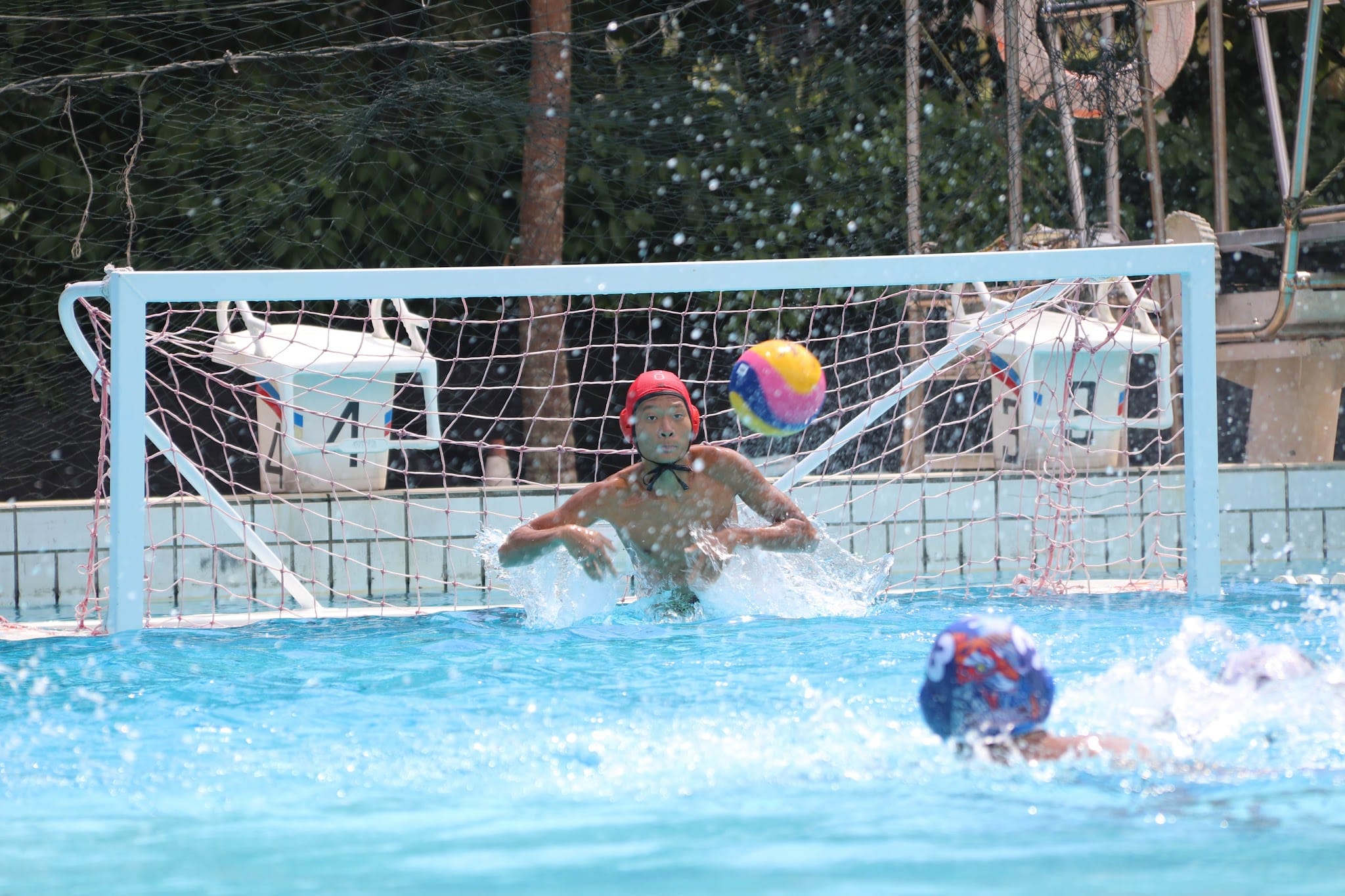 A water polo goalkeeper in a red cap defends the goal as a colorful ball flies toward the net.