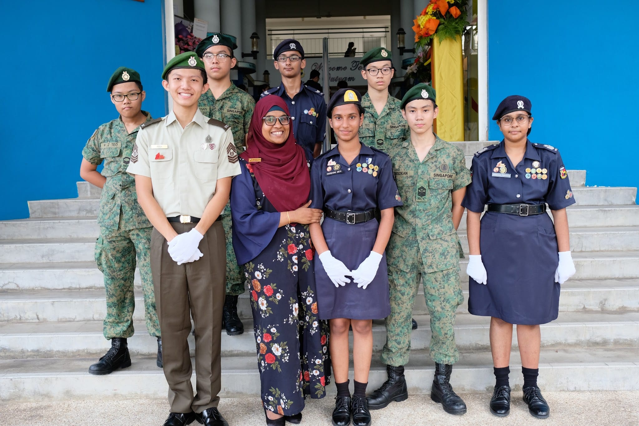 A group of young cadets in military and navy uniforms pose with a woman in a floral dress on building steps.
