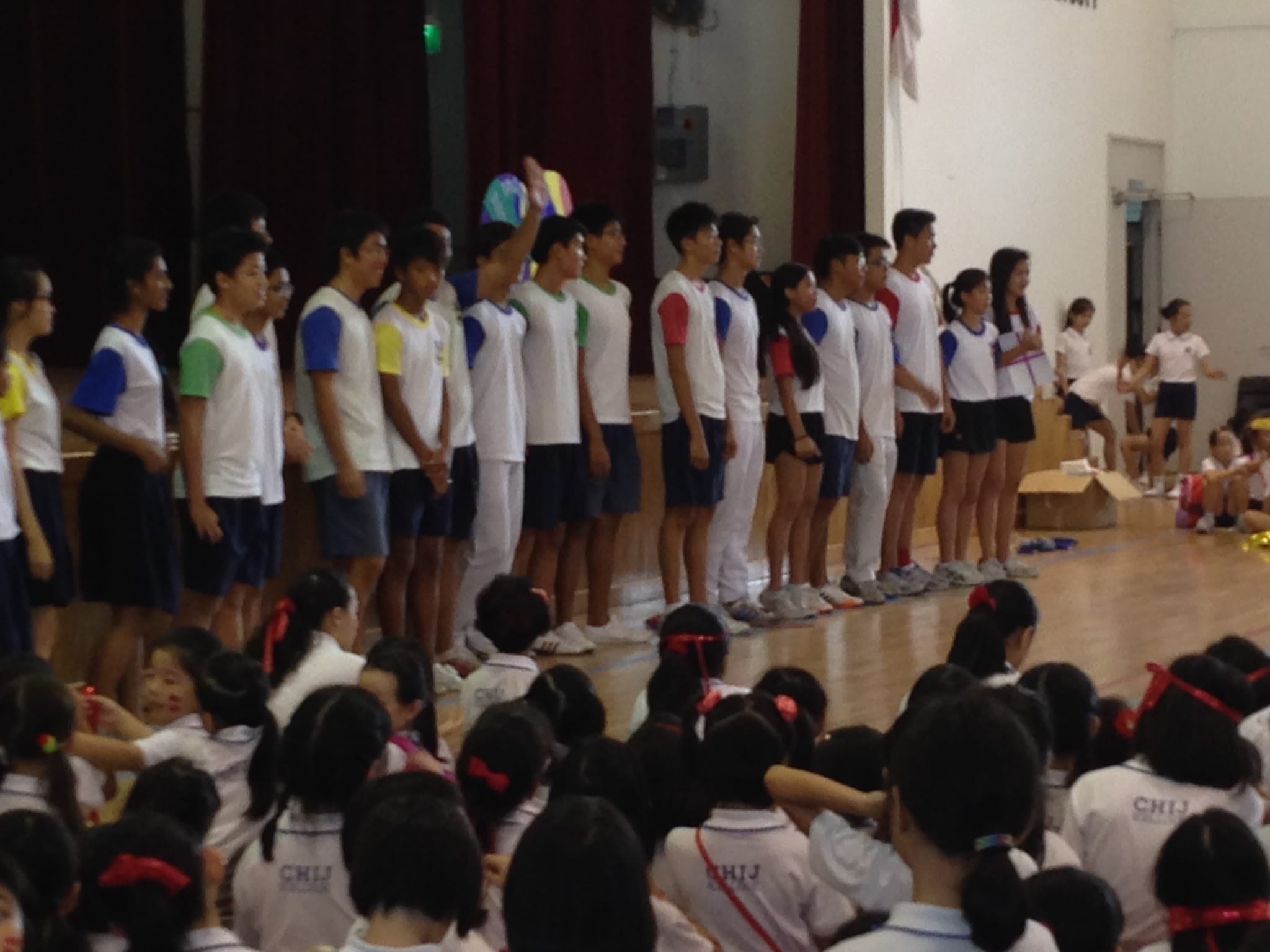 Students in colorful sports jerseys stand in a line on a gymnasium floor while seated students in white uniforms watch.