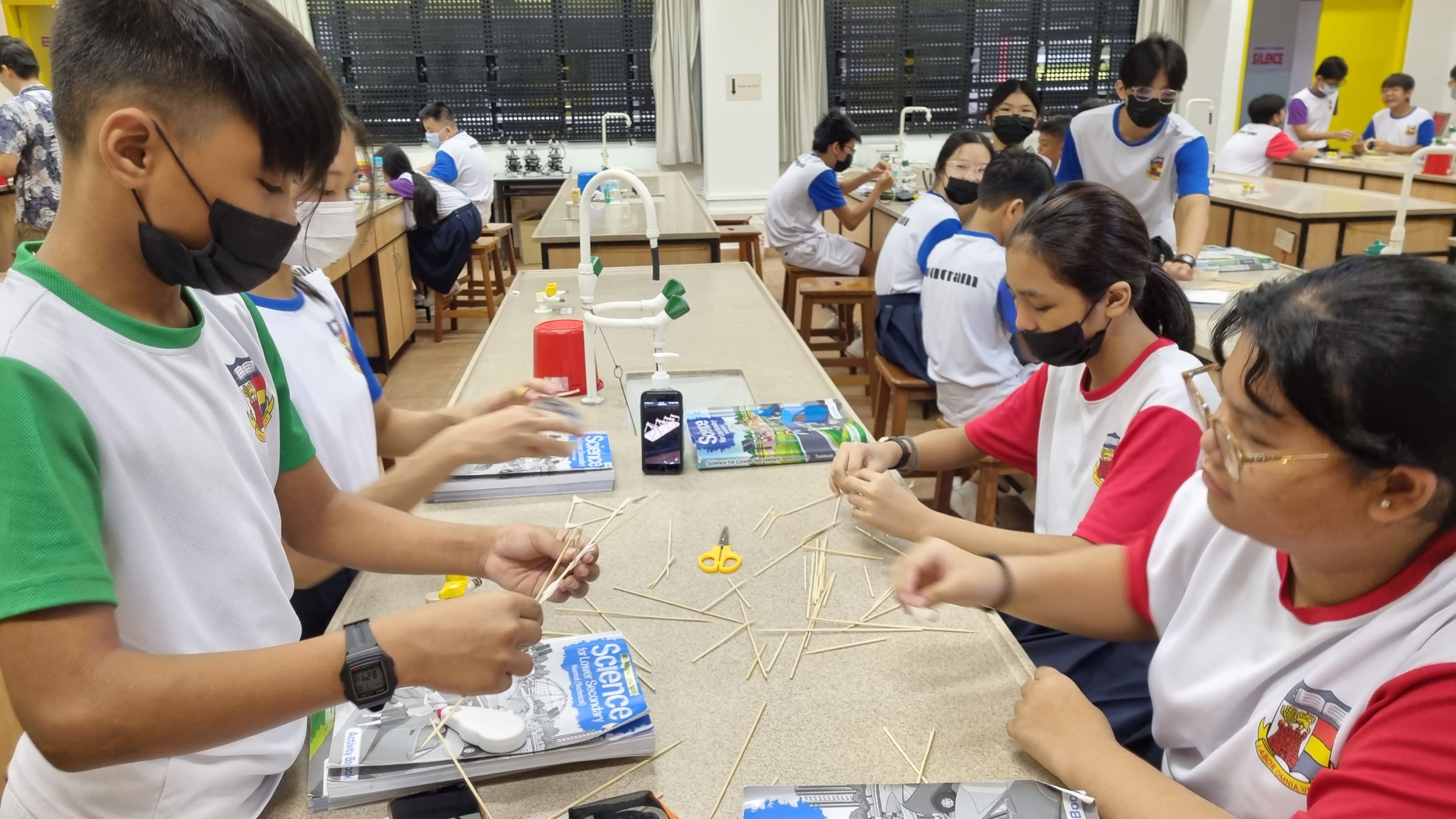 Students wearing masks work with wooden sticks and scissors on lab benches in a school science classroom.