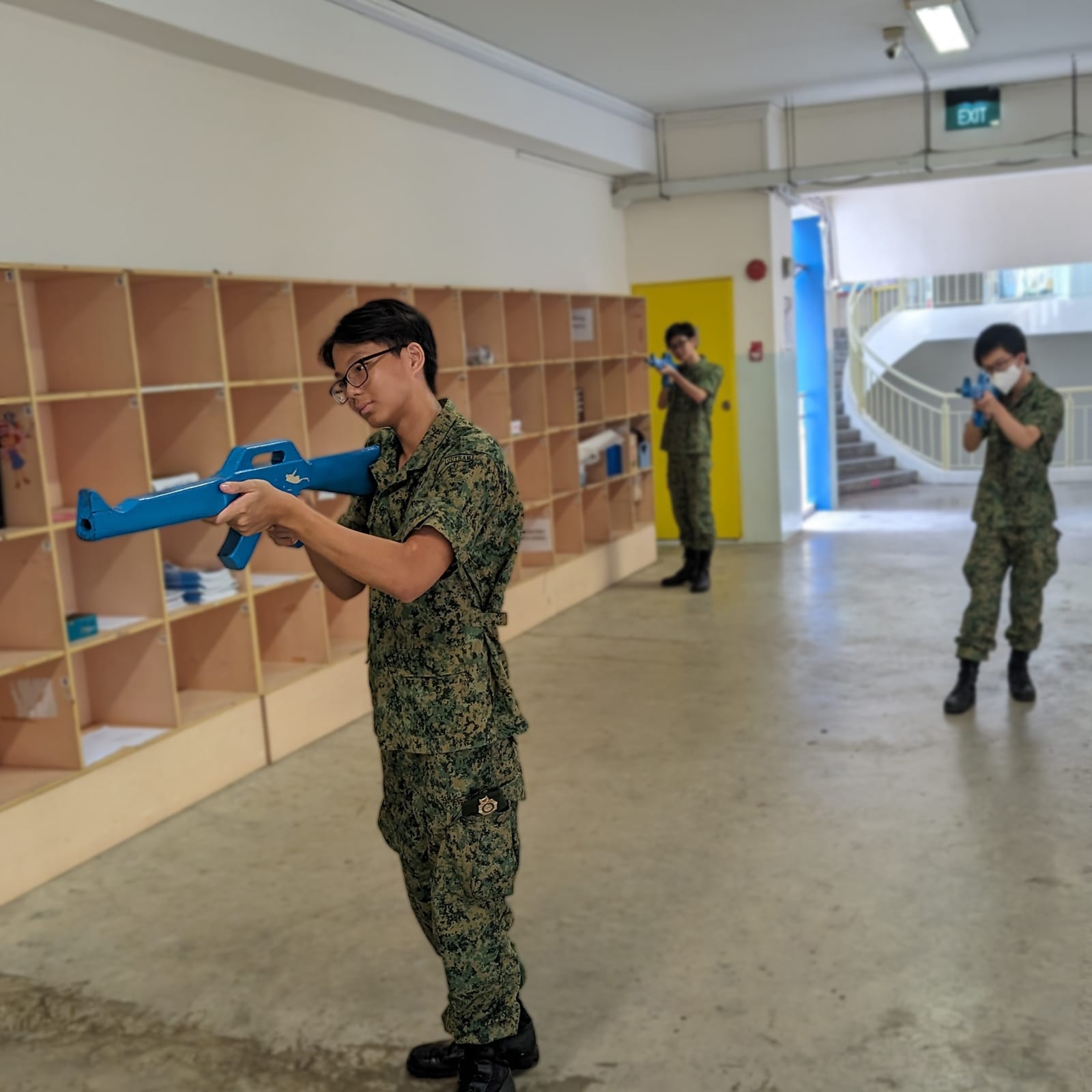 A cadet in camouflage uniform aims a blue training rifle down a corridor, with two more cadets aiming similarly in the background.