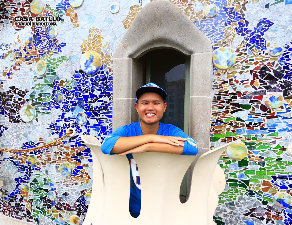 Young man in a blue shirt and cap smiling at Casa Batlló, leaning on a balcony surrounded by colorful mosaic tiles.