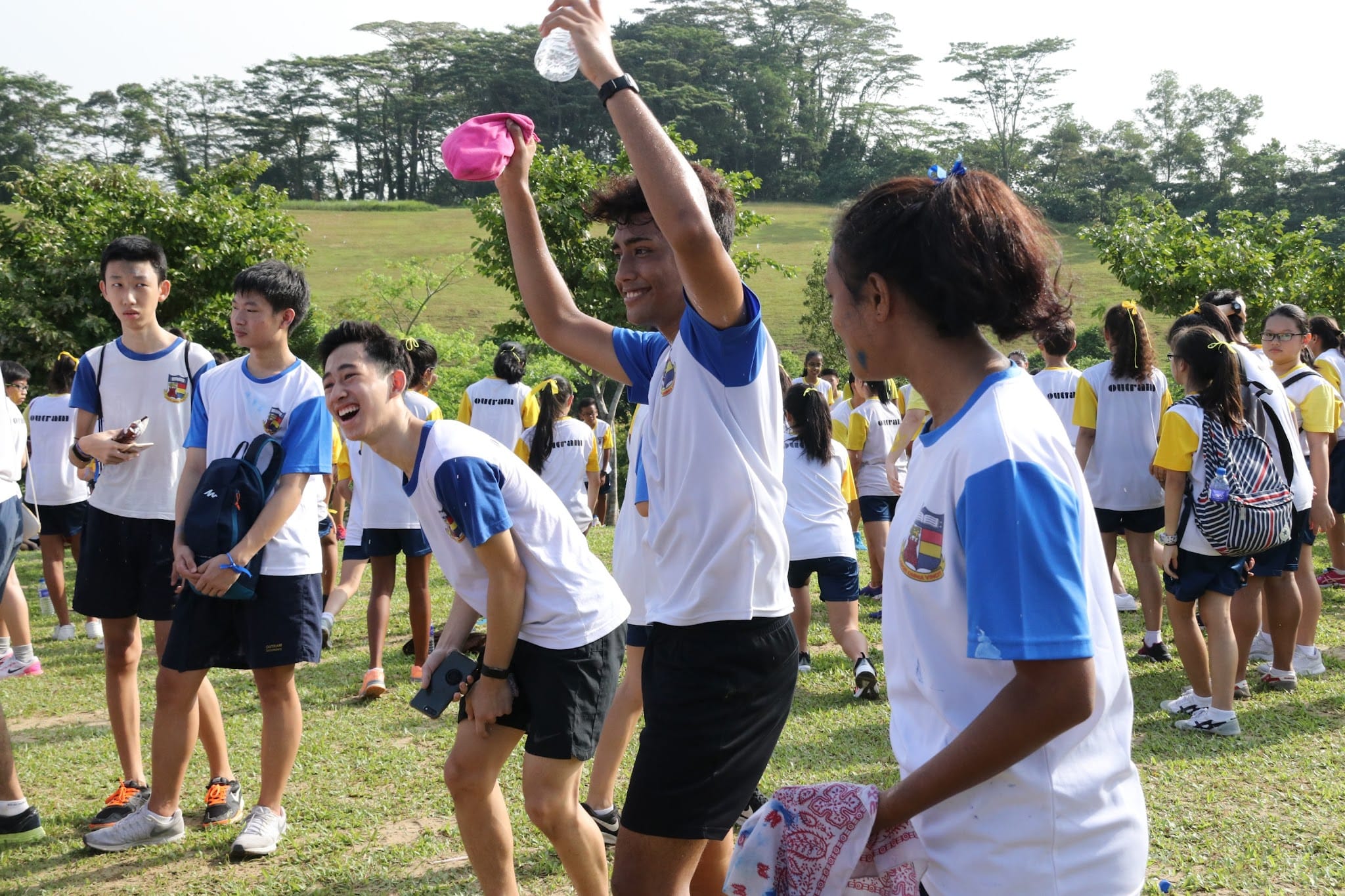 Students in white and blue school uniforms gather on a grassy field outdoors, one boy raising a water bottle and smiling.
