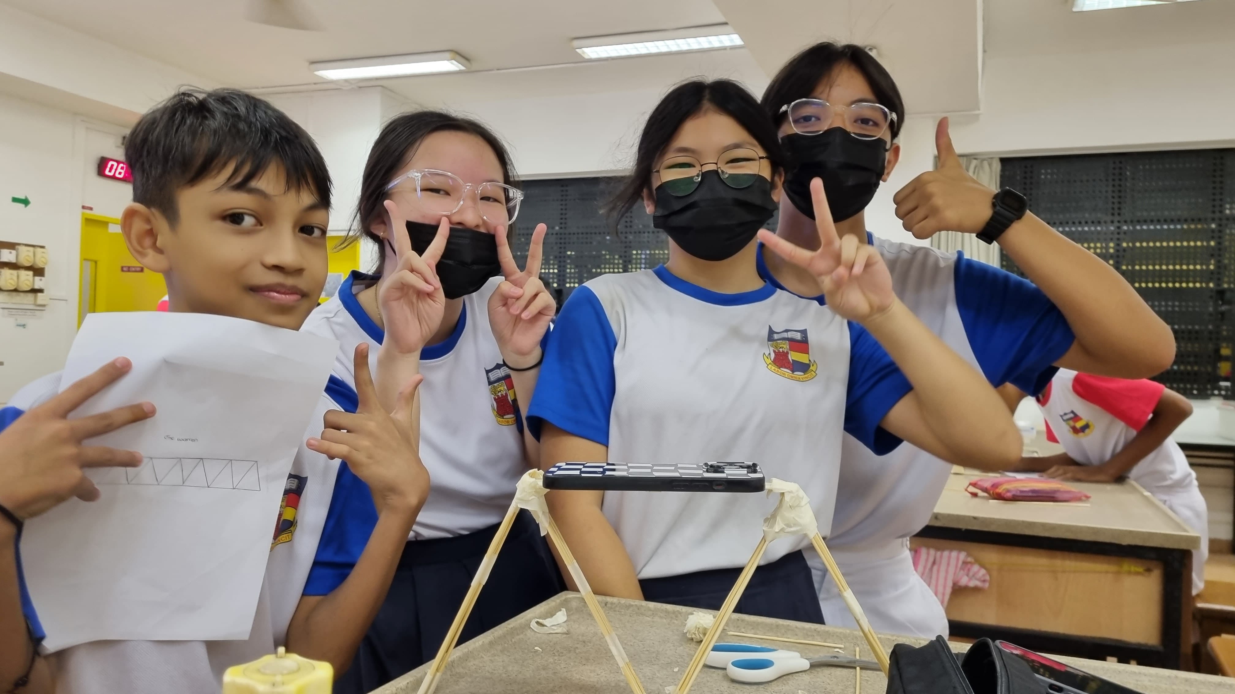 Four students in school uniforms pose with peace signs near a wooden stick structure on a desk in a classroom.