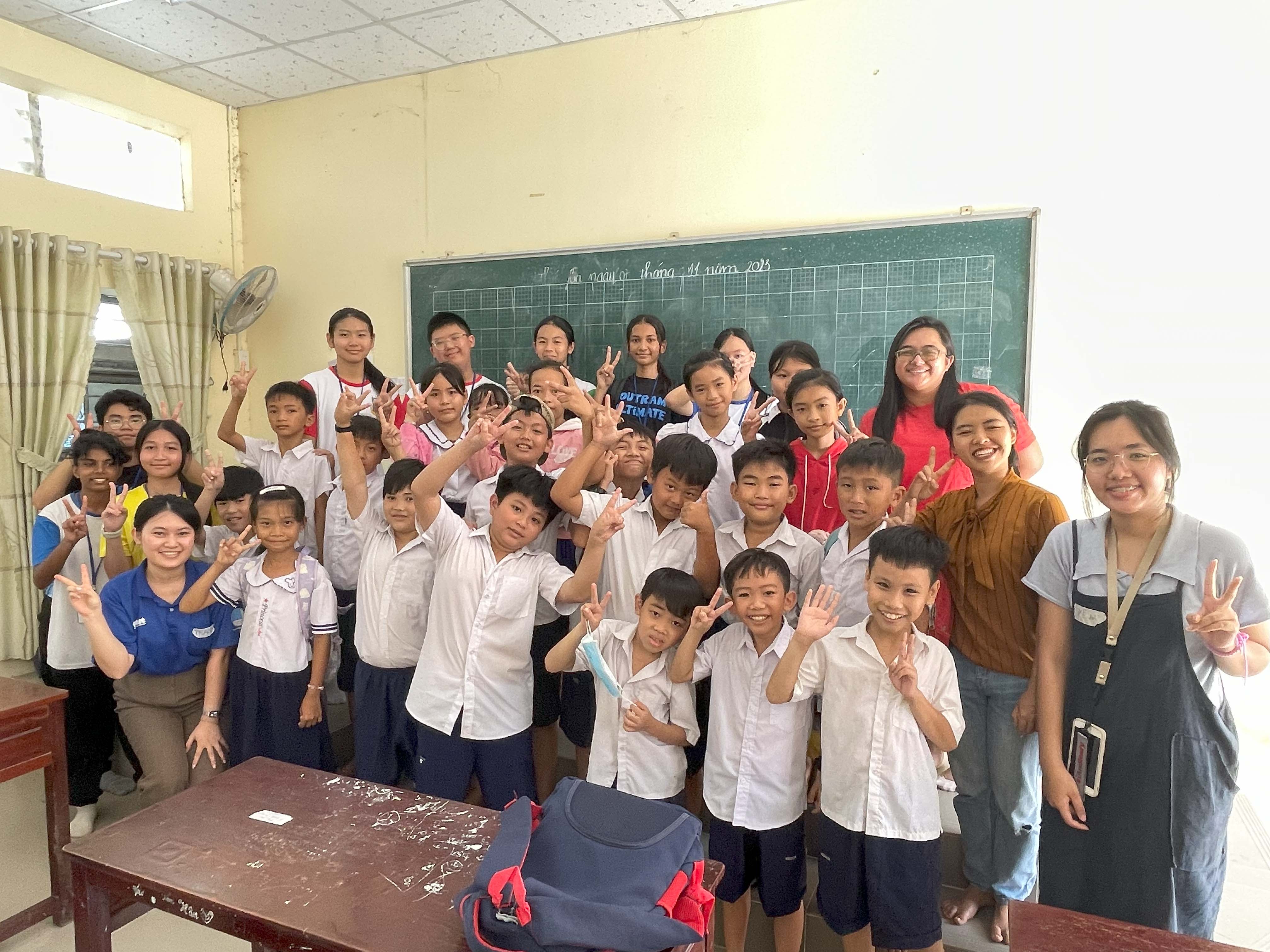 A large group of smiling children in white uniforms and several adults pose with peace signs in a classroom.