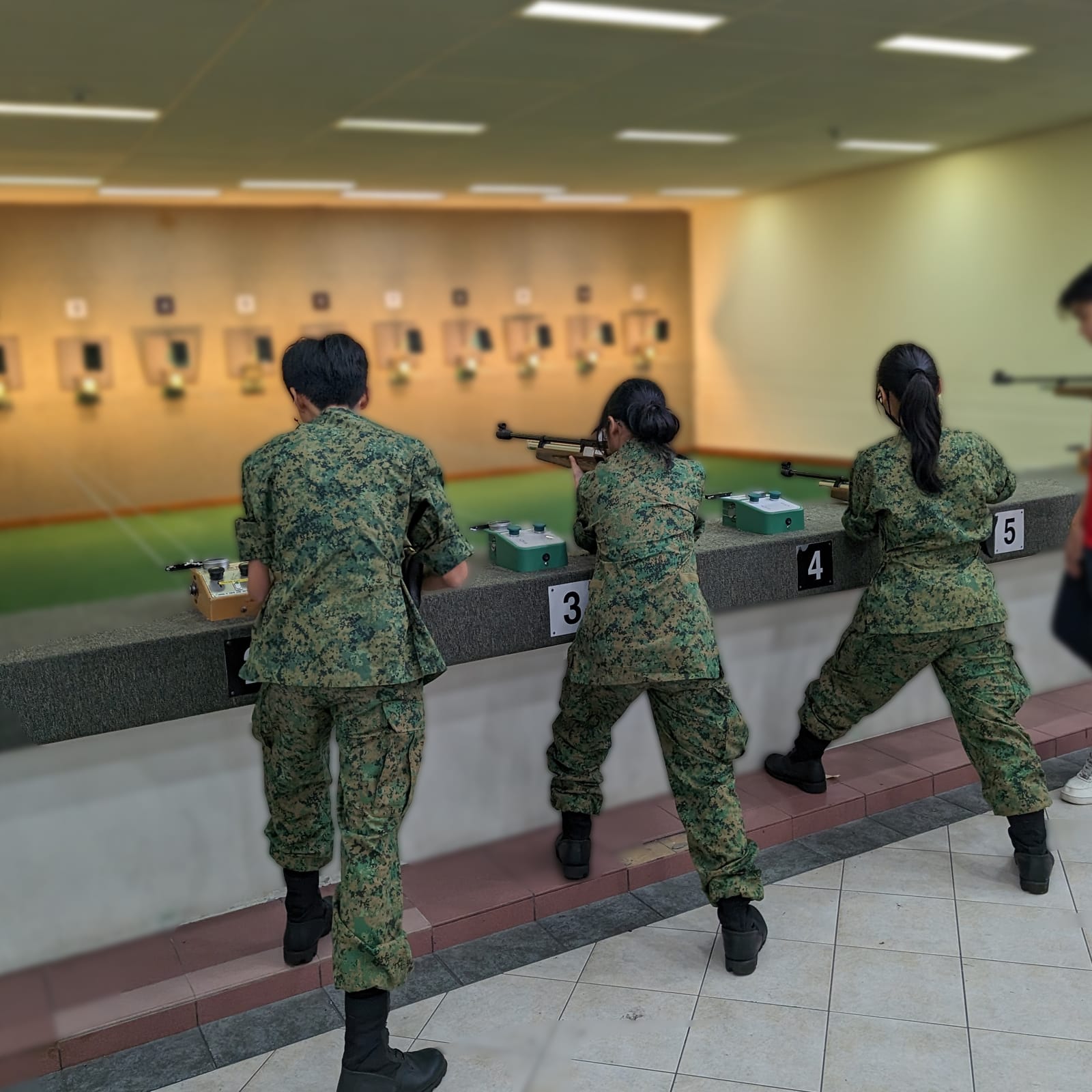 Three cadets in camouflage uniform aim air rifles at targets on an indoor shooting range, standing at numbered firing points 3, 4, and 5.