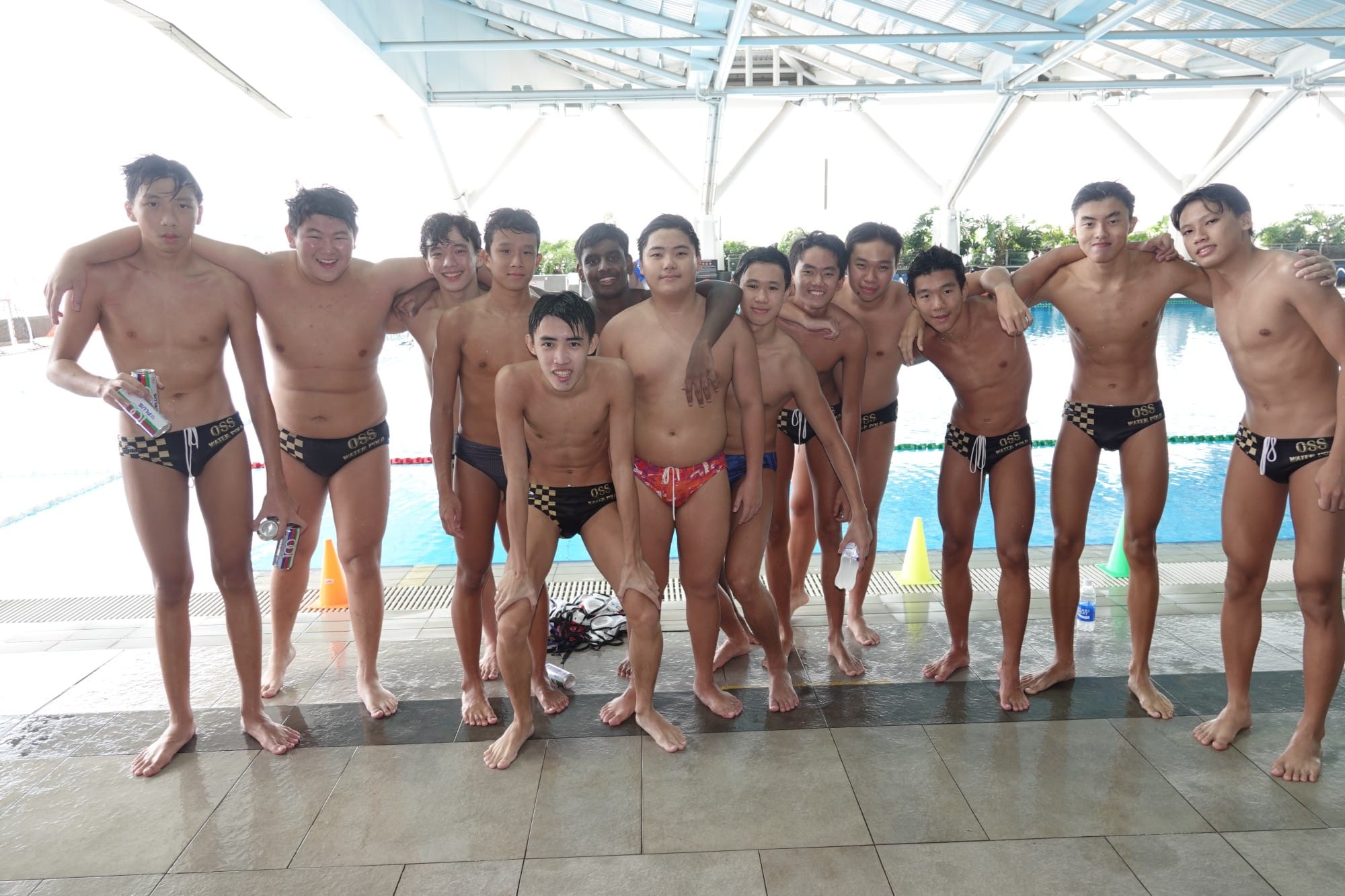 A group of young male water polo players in OSS swimwear posing together beside an indoor pool.