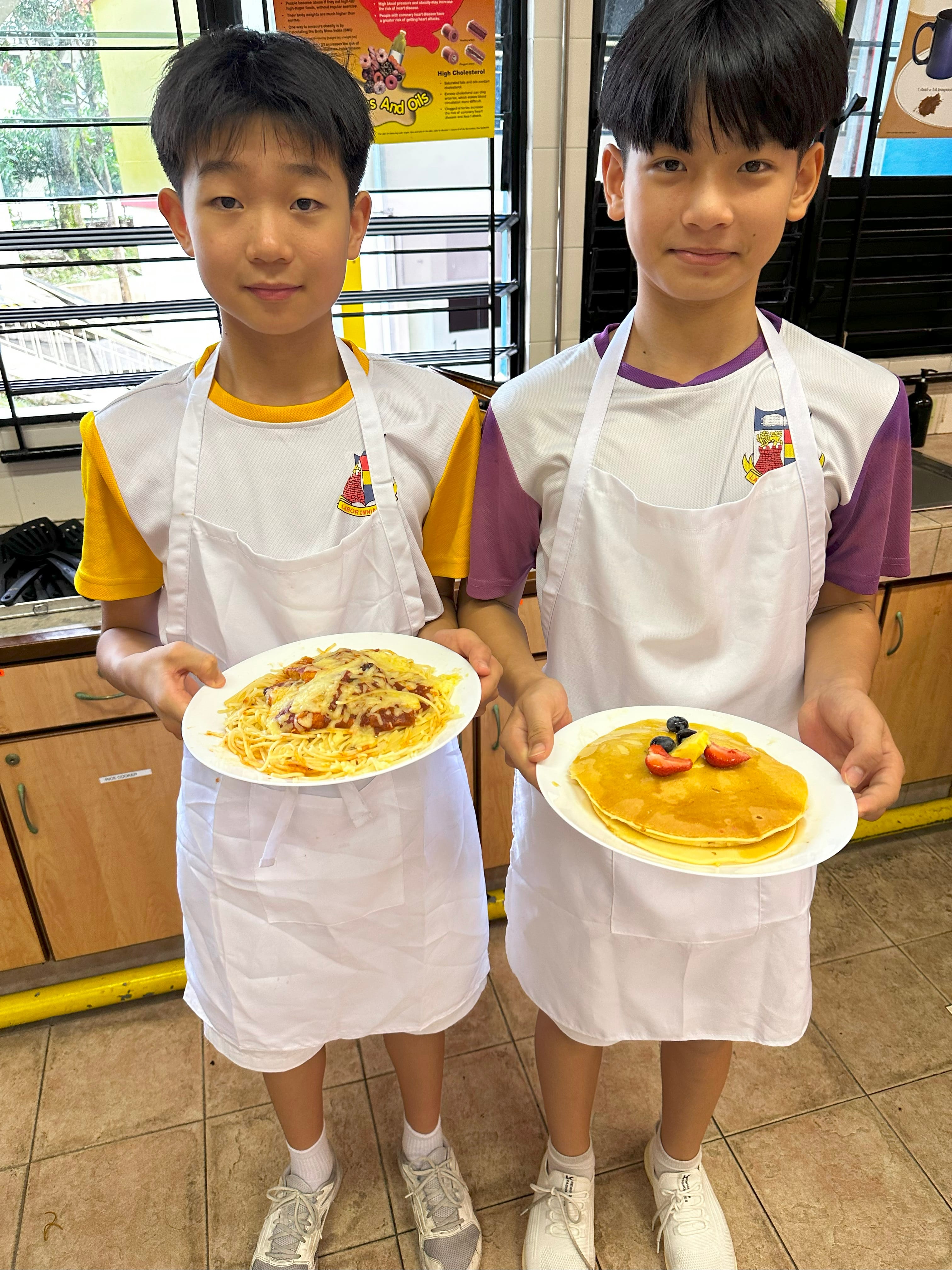 Two boys in white aprons hold plates of spaghetti with sauce and pancakes topped with fruit.
