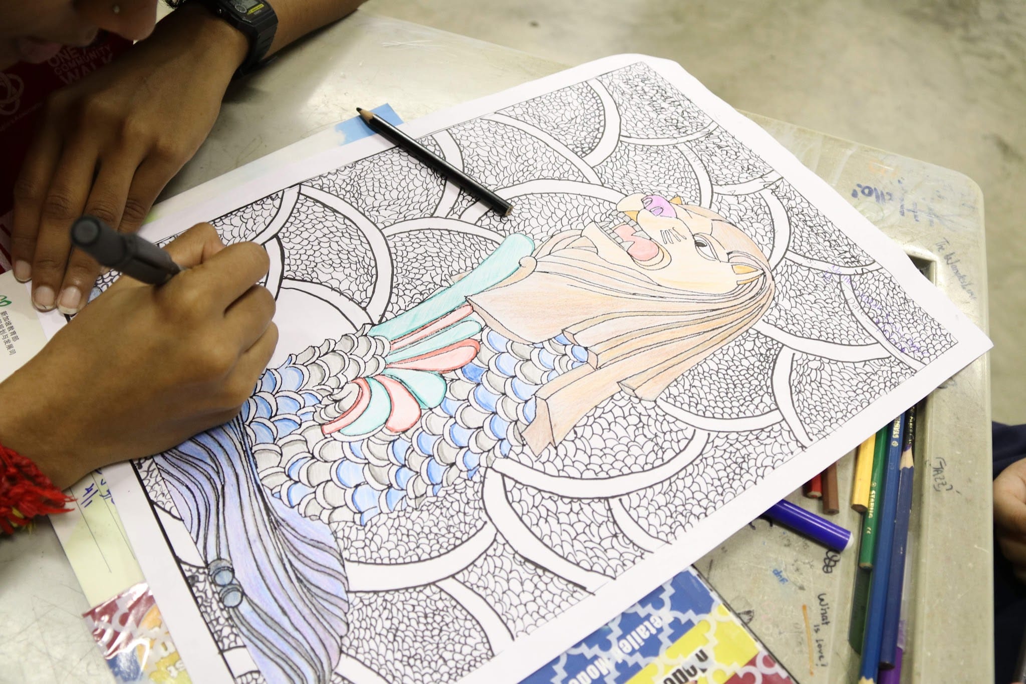 A person colors a detailed anime mermaid drawing with colored pencils on a desk.