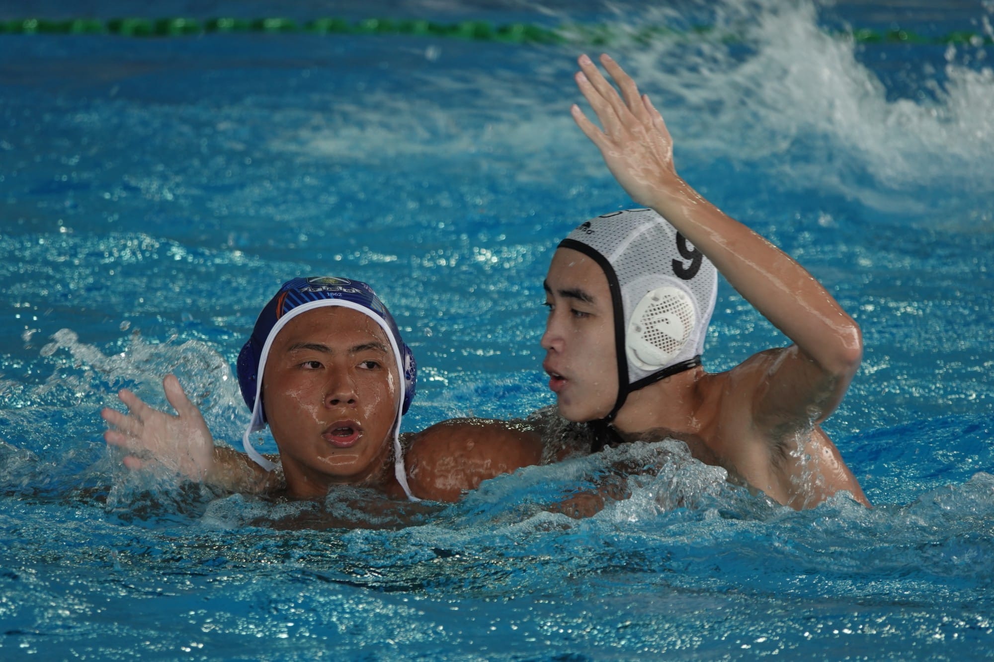 Two male water polo players in a pool, one in a blue cap and one in a white cap numbered 9, competing closely.