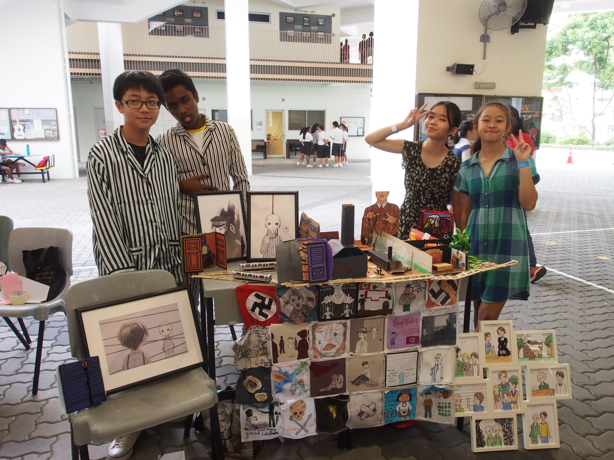 Four students stand beside a display table covered with hand-drawn artwork and framed illustrations at a school event.