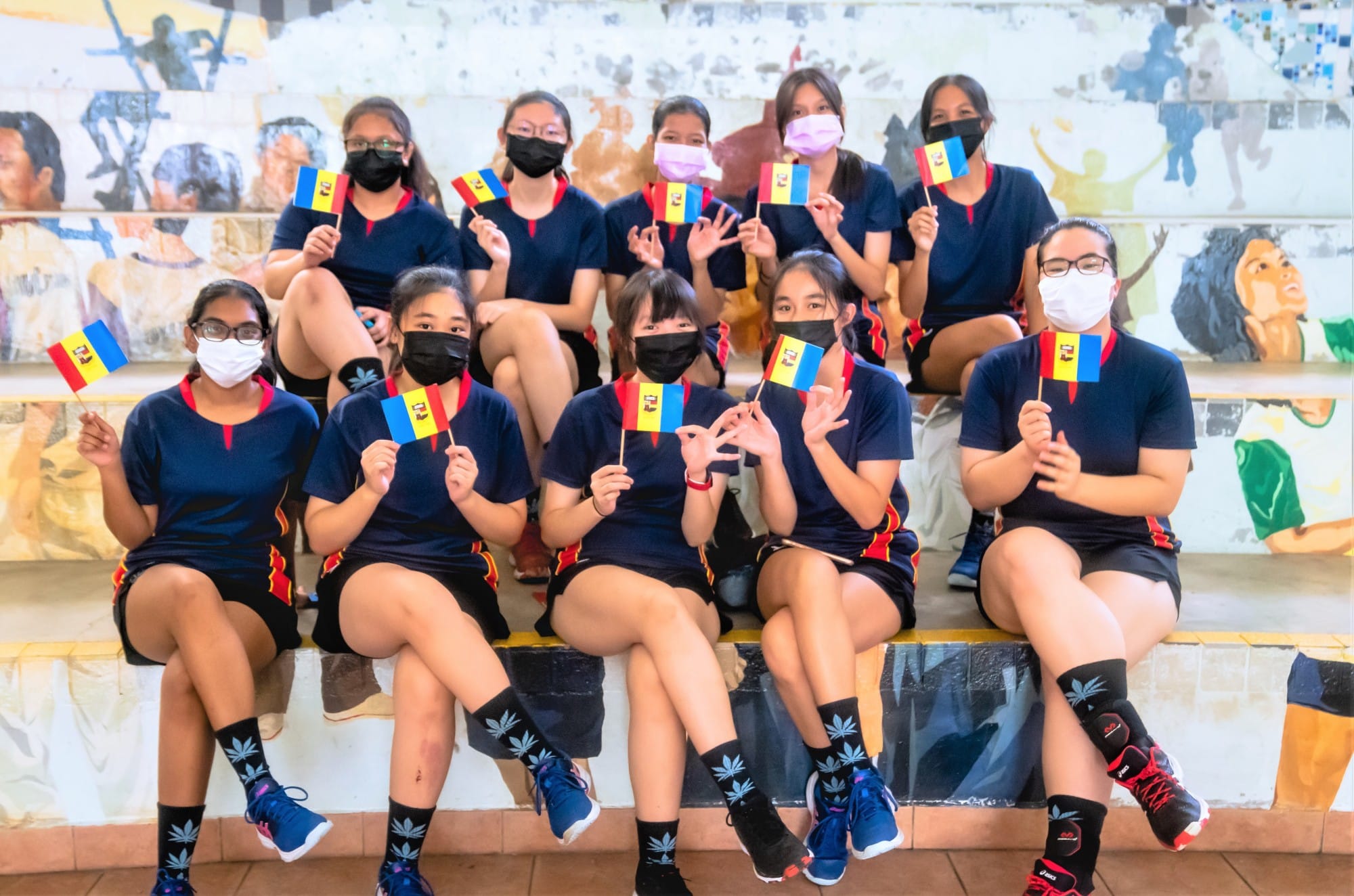 A group of masked girls in navy uniforms sit on steps holding small Venezuelan flags in front of a colorful mural.