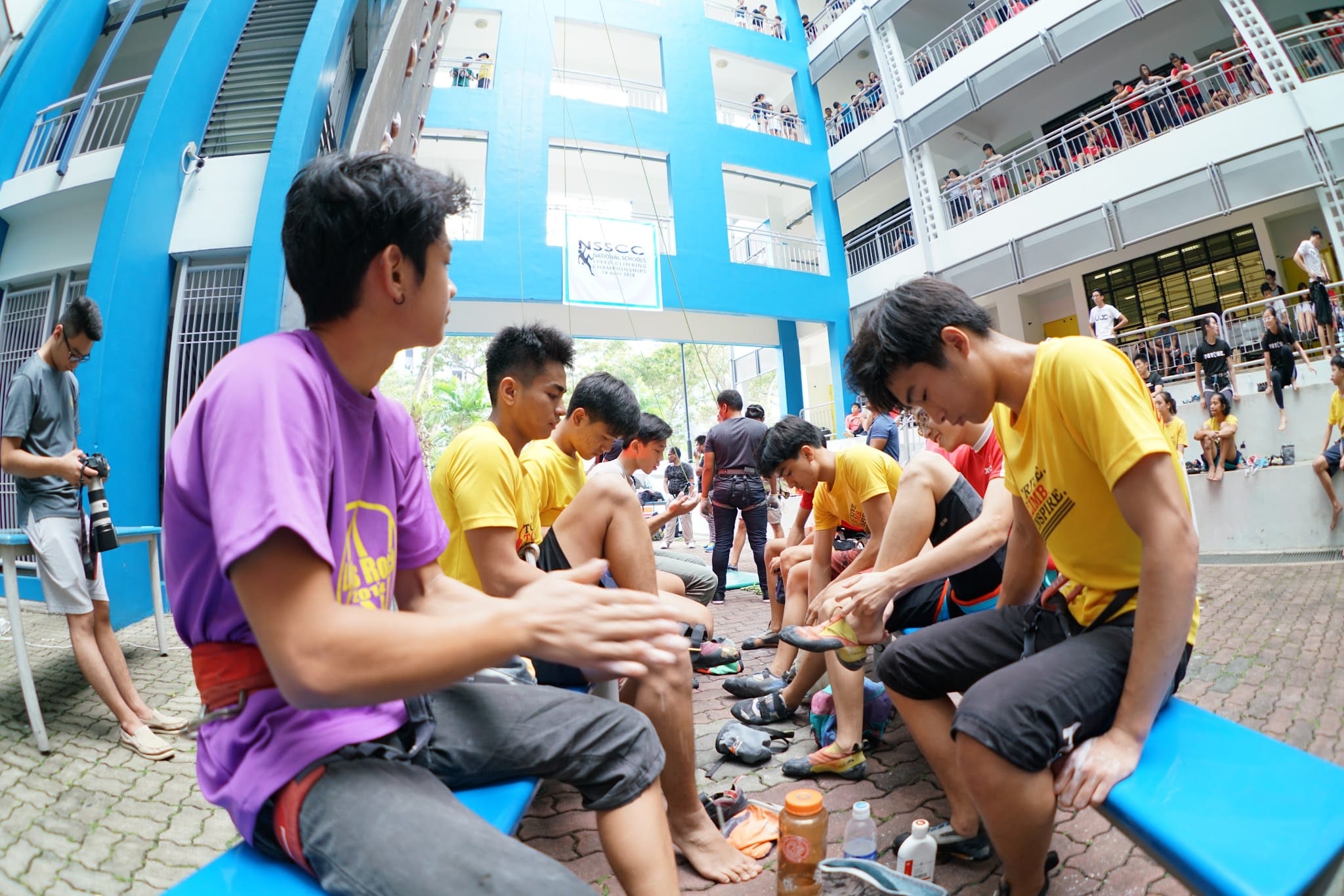 Young athletes in yellow and purple shirts put on climbing shoes near a blue climbing wall at an outdoor competition.
