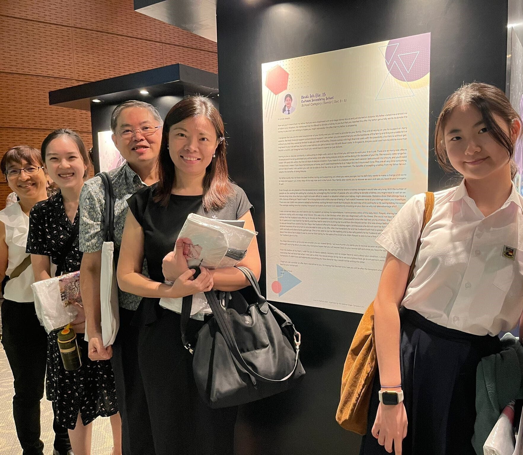 Four people smile beside a large poster display at an indoor exhibition with brick walls.
