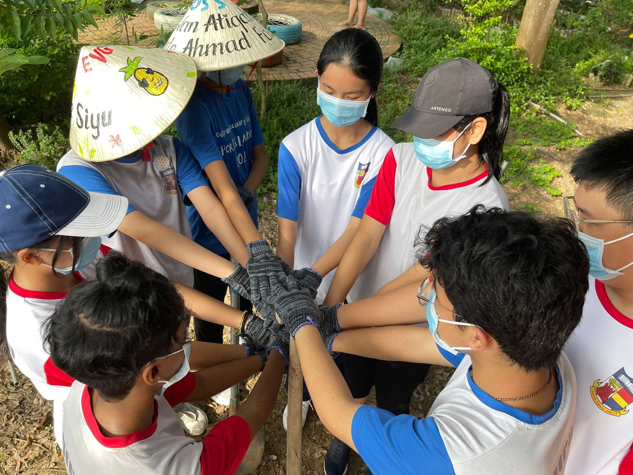 A group of masked youth wearing matching uniforms stack their gloved hands together in a team huddle outdoors.