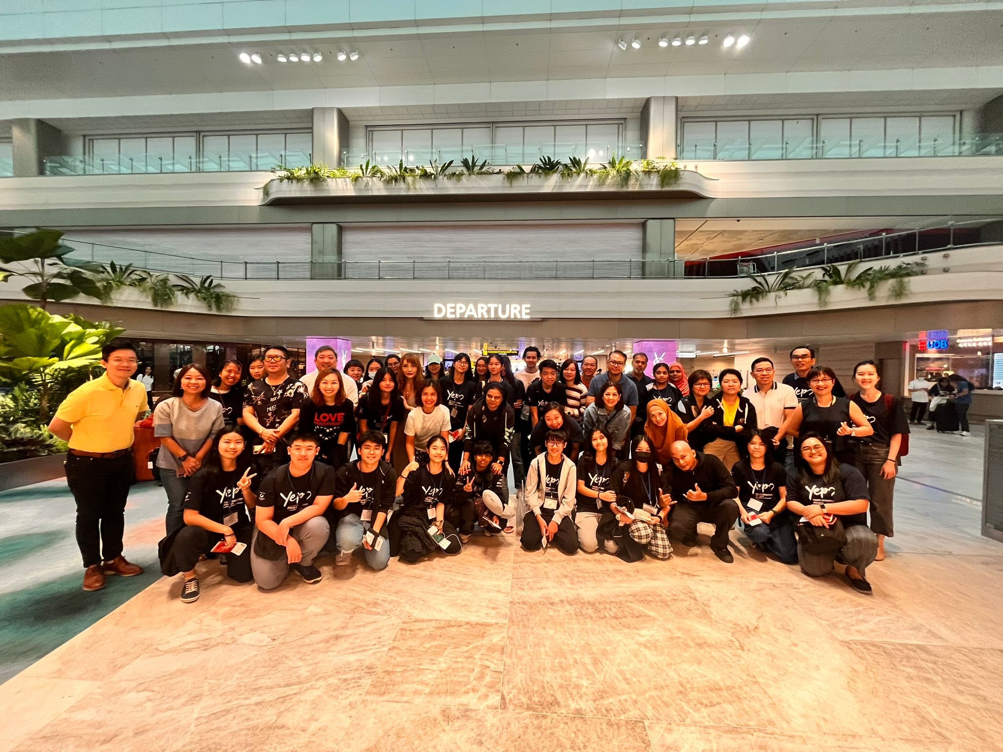 Large group of people posing together in front of a Departure sign at an airport terminal.