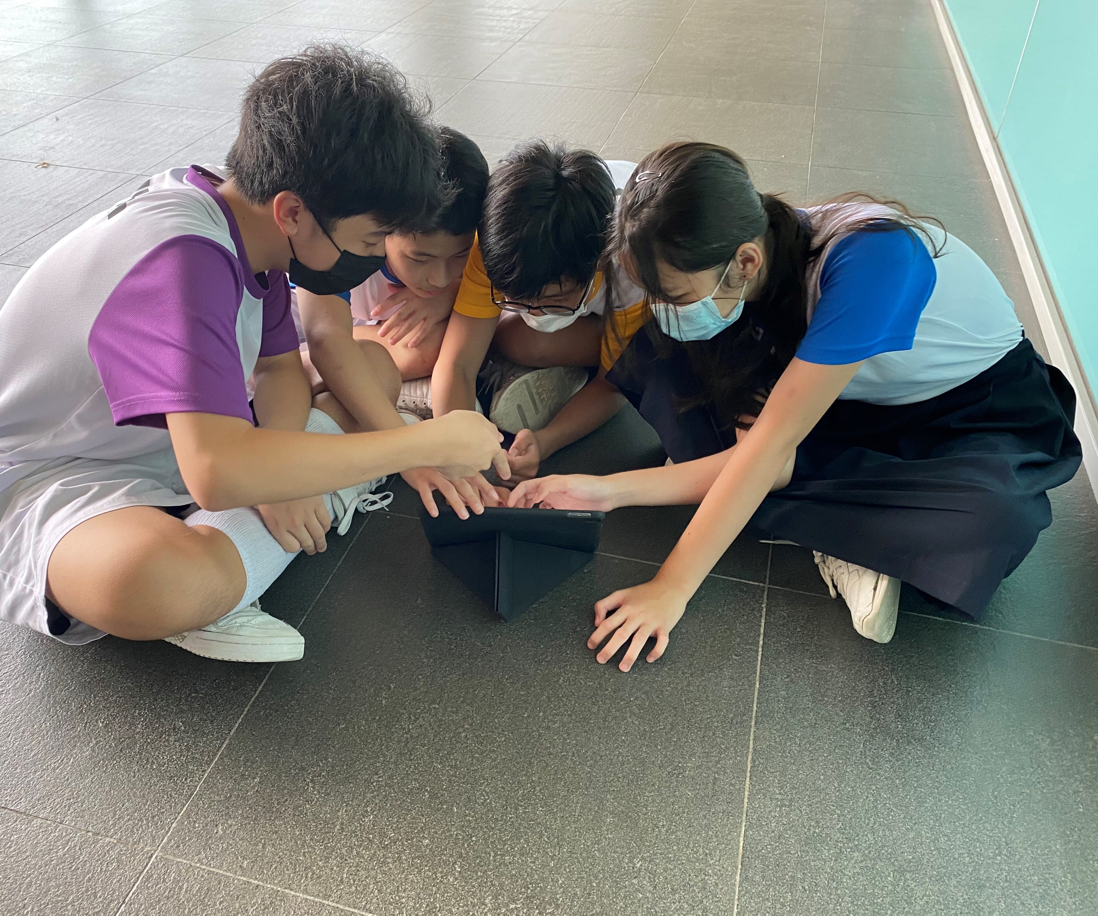 Four masked students in school uniforms sit on a tiled floor, huddled together tapping on a tablet.
