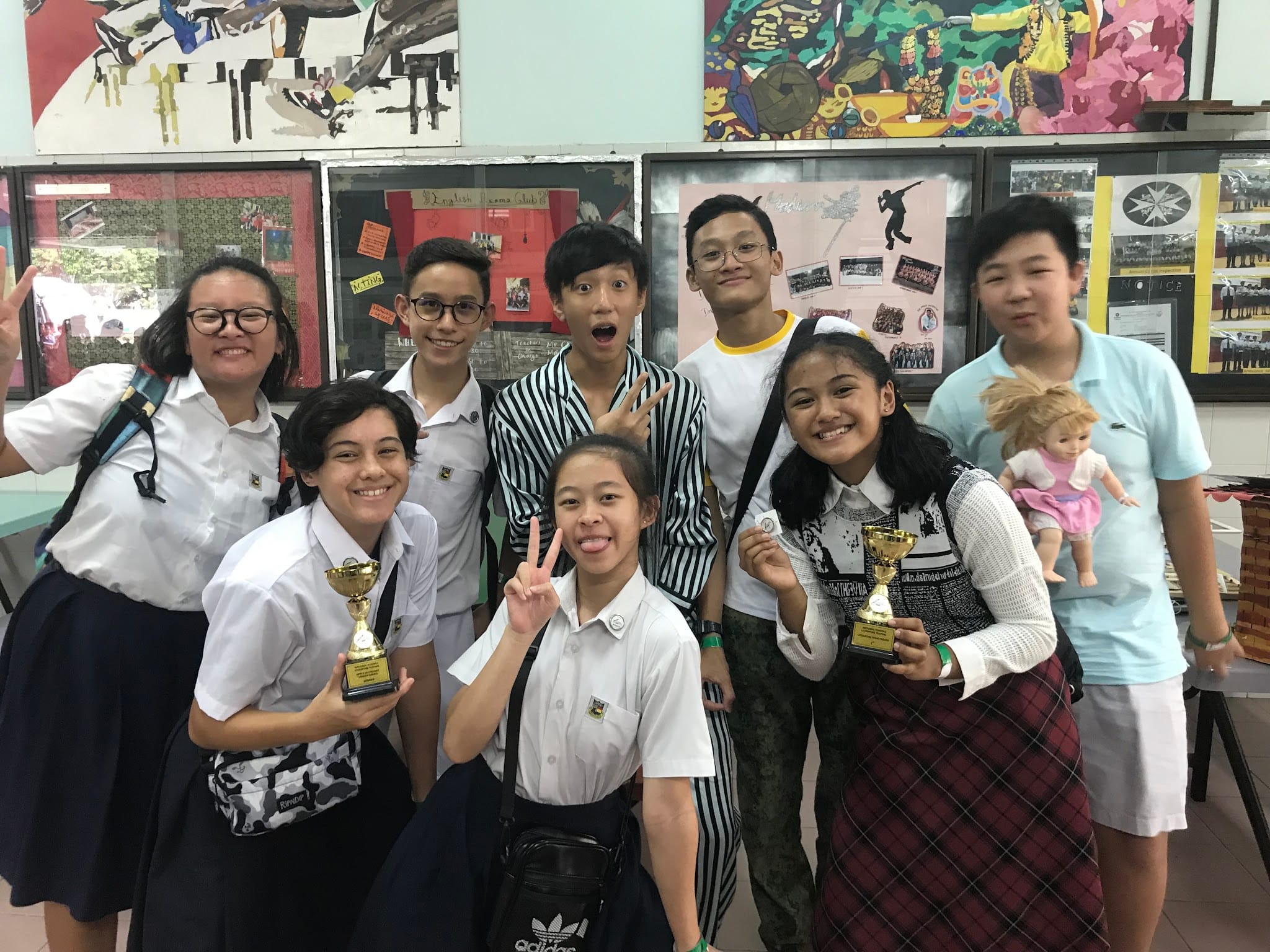 Eight smiling students in school uniforms pose together, two holding small gold trophies, in front of colorful wall displays.