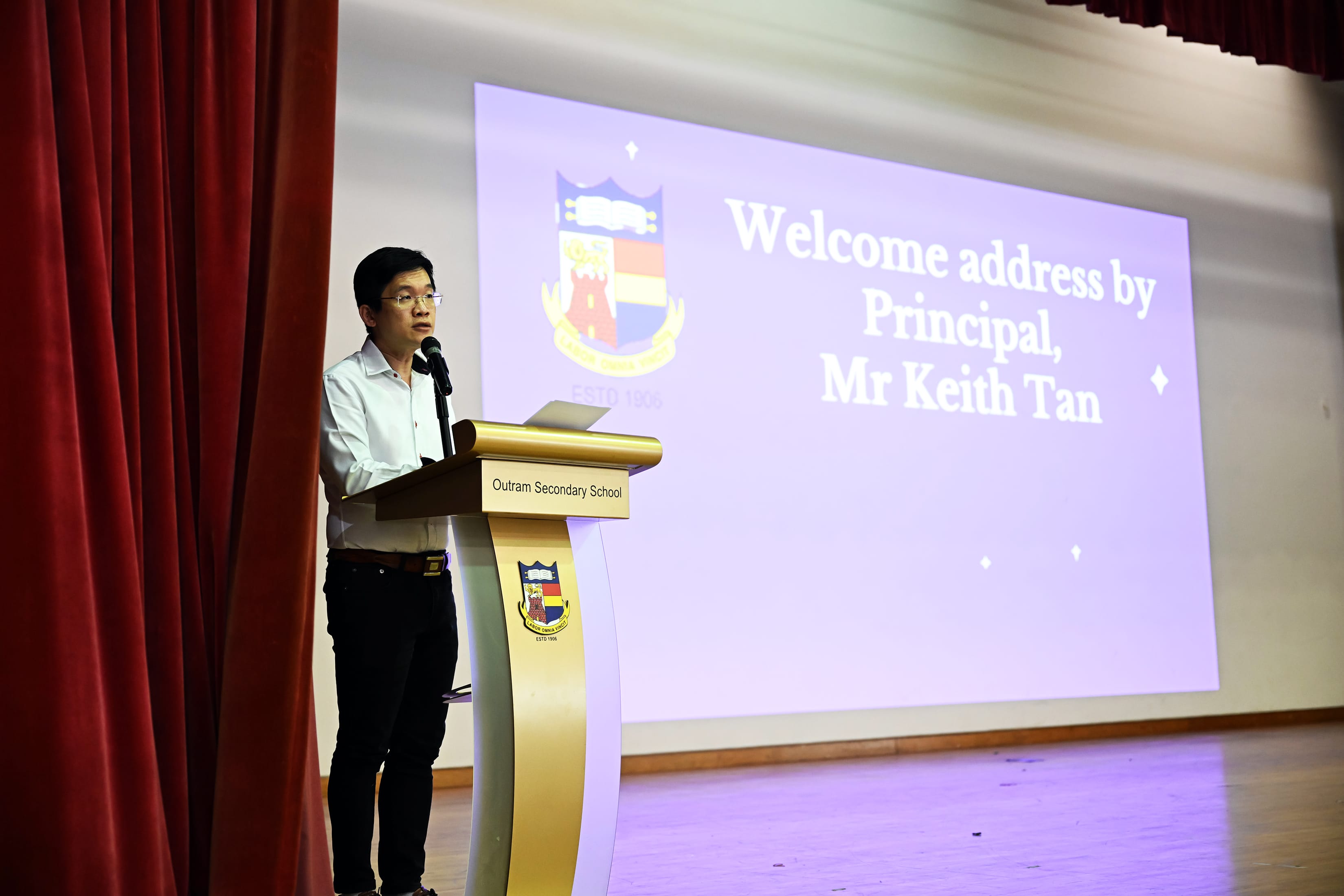 Man speaking at Outram Secondary School podium, with a projected slide reading "Welcome address by Principal, Mr Keith Tan."