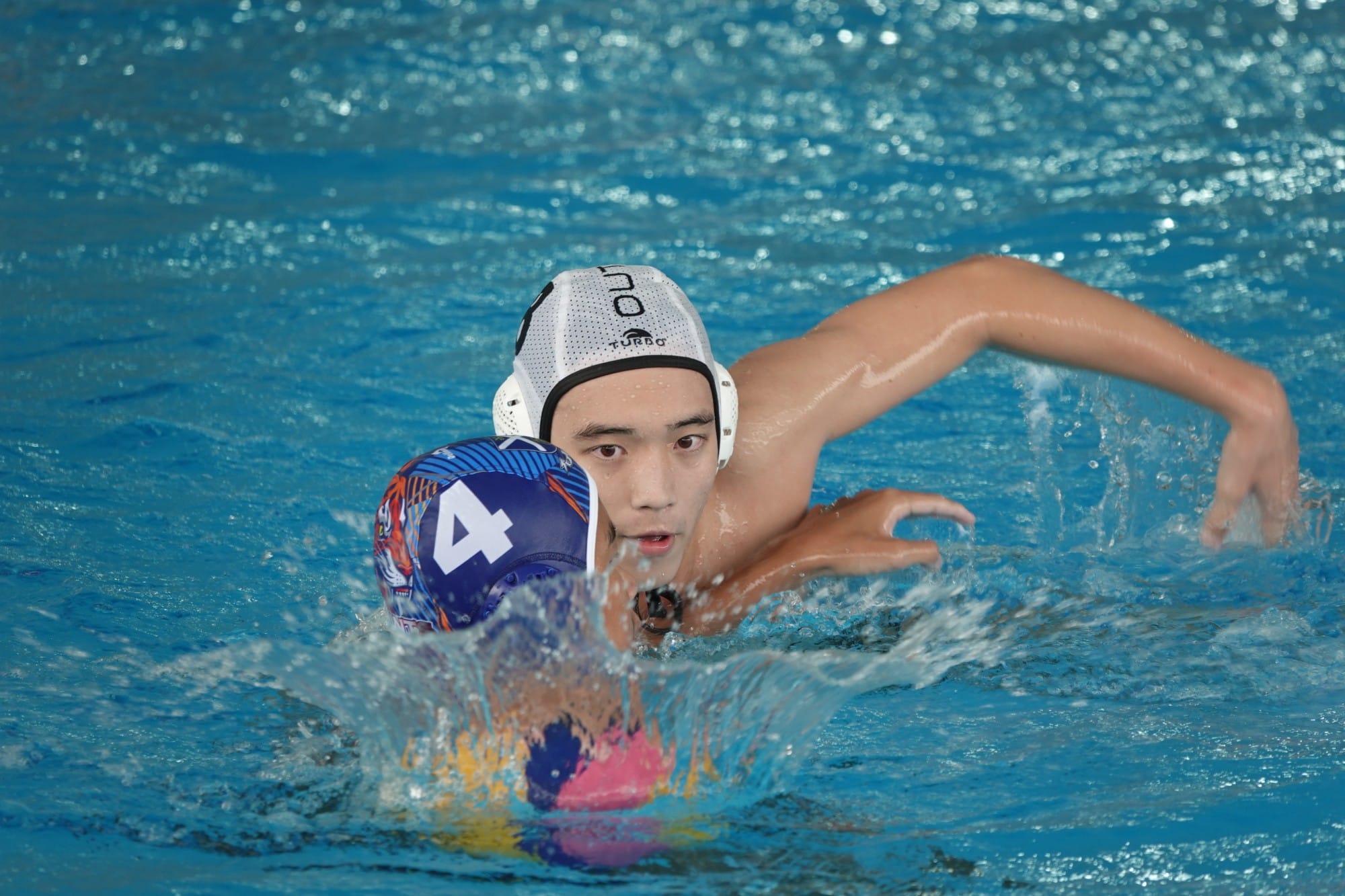 A young male water polo player in a white cap reaches for a ball near a player wearing a blue number 4 cap.