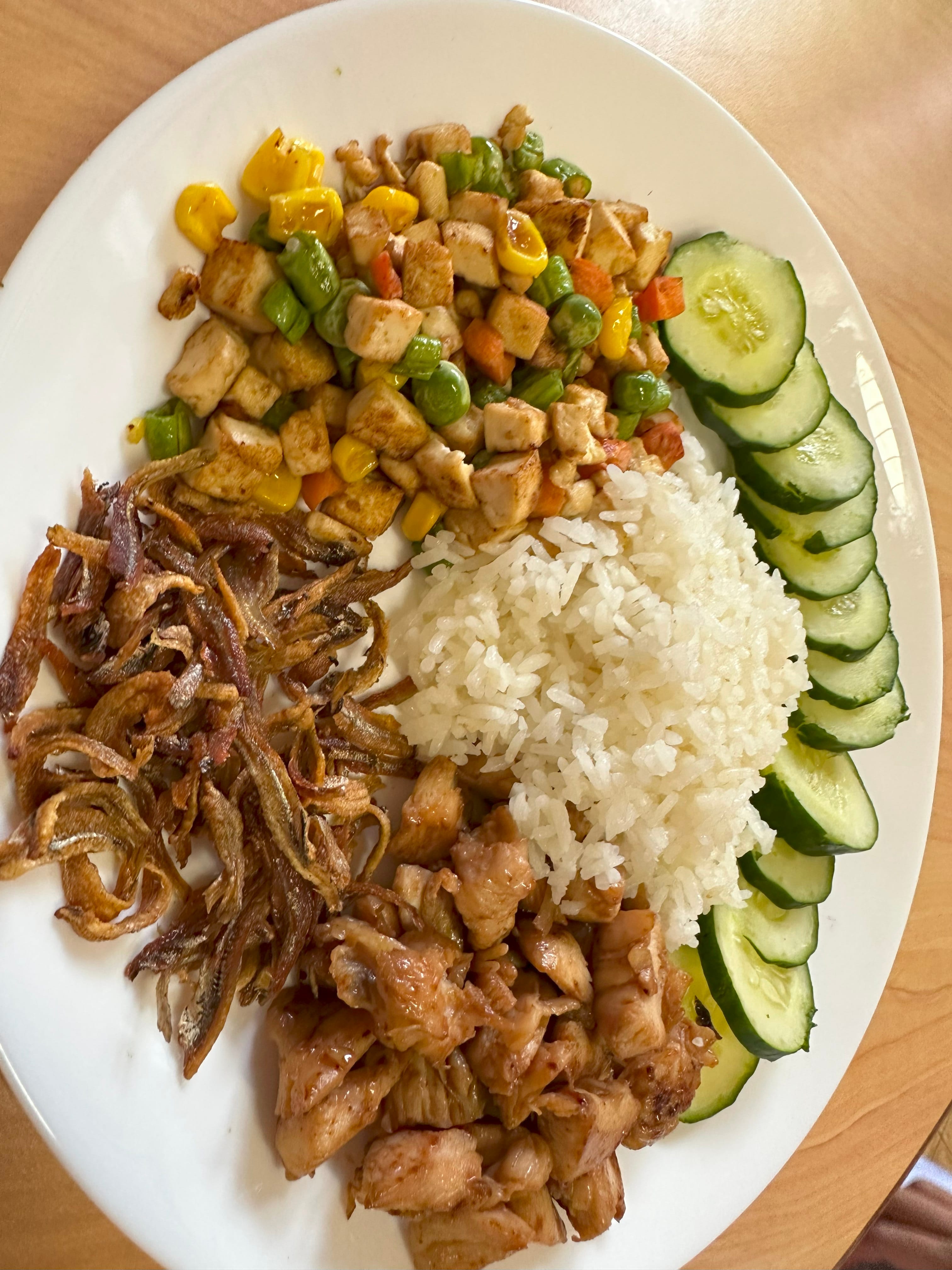 White plate with rice, grilled chicken, stir-fried vegetables, fried anchovies, and sliced cucumbers arranged around it.