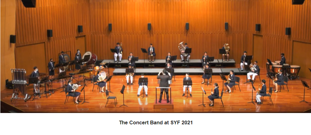 A concert band with students in uniform performing on a wooden stage with a conductor at SYF 2021.