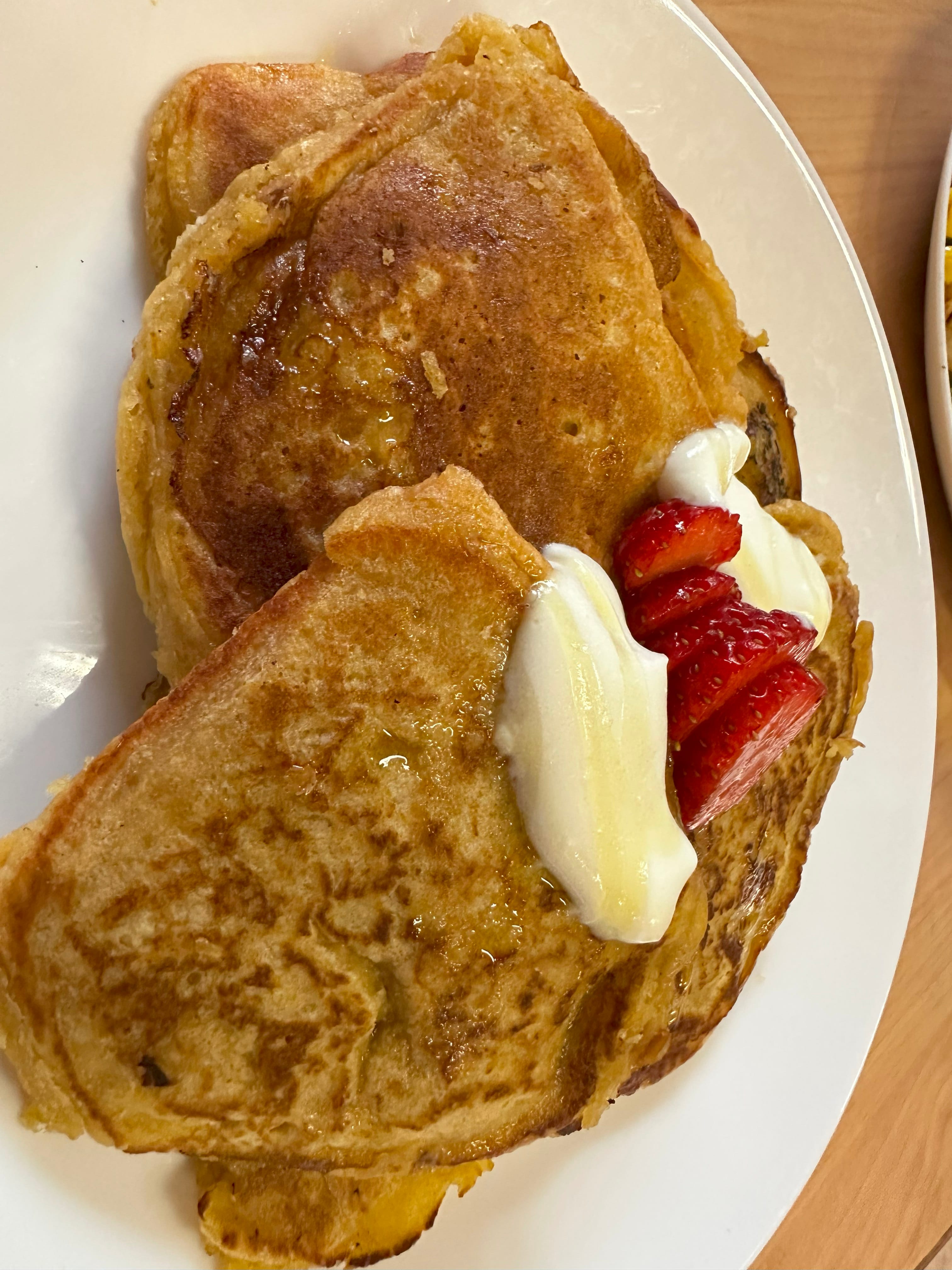 Golden pancakes on a white plate topped with whipped cream, sliced strawberries, and syrup.