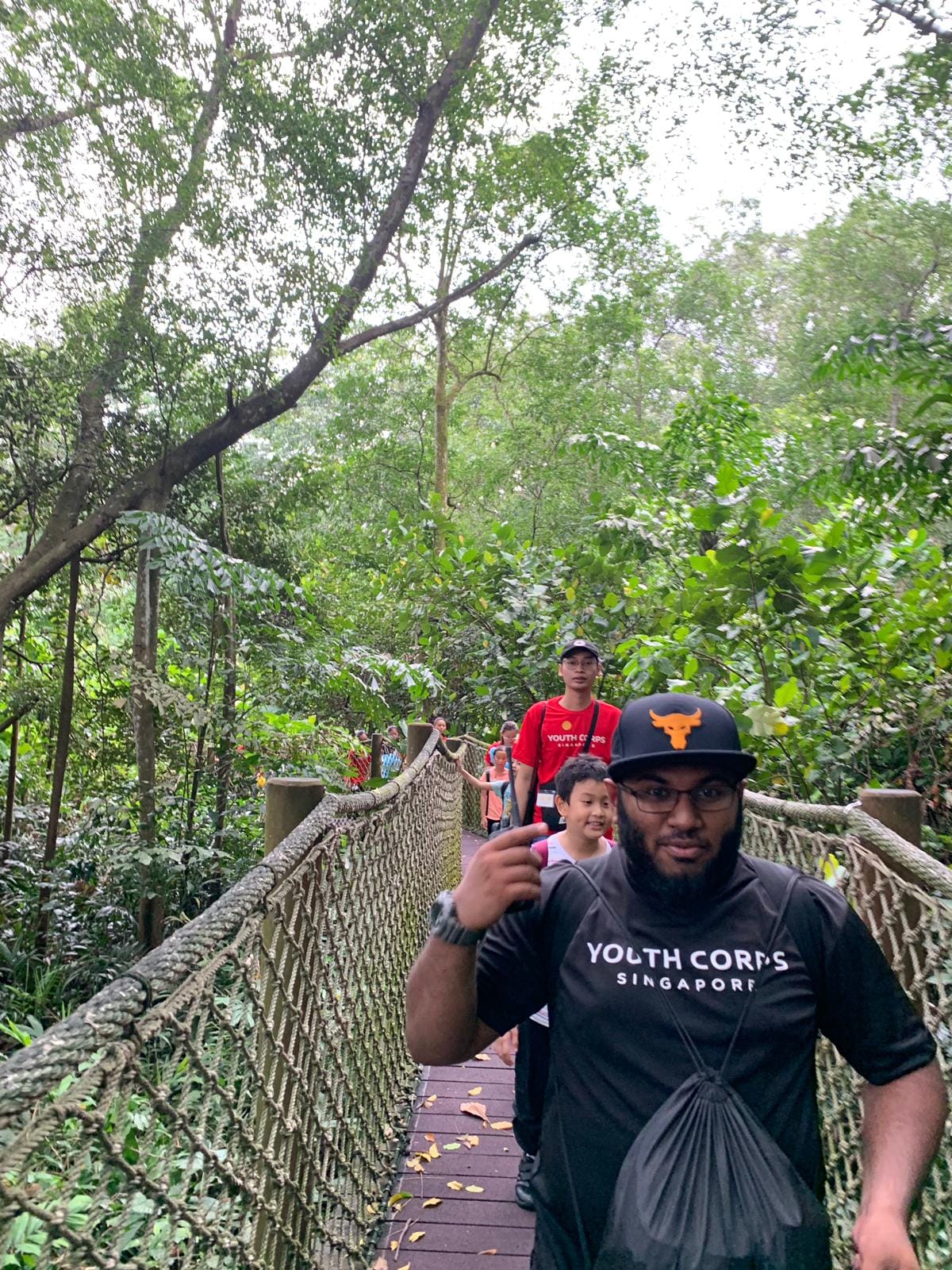 A group wearing Youth Corps Singapore shirts walks across a rope-railed wooden suspension bridge through a lush forest.