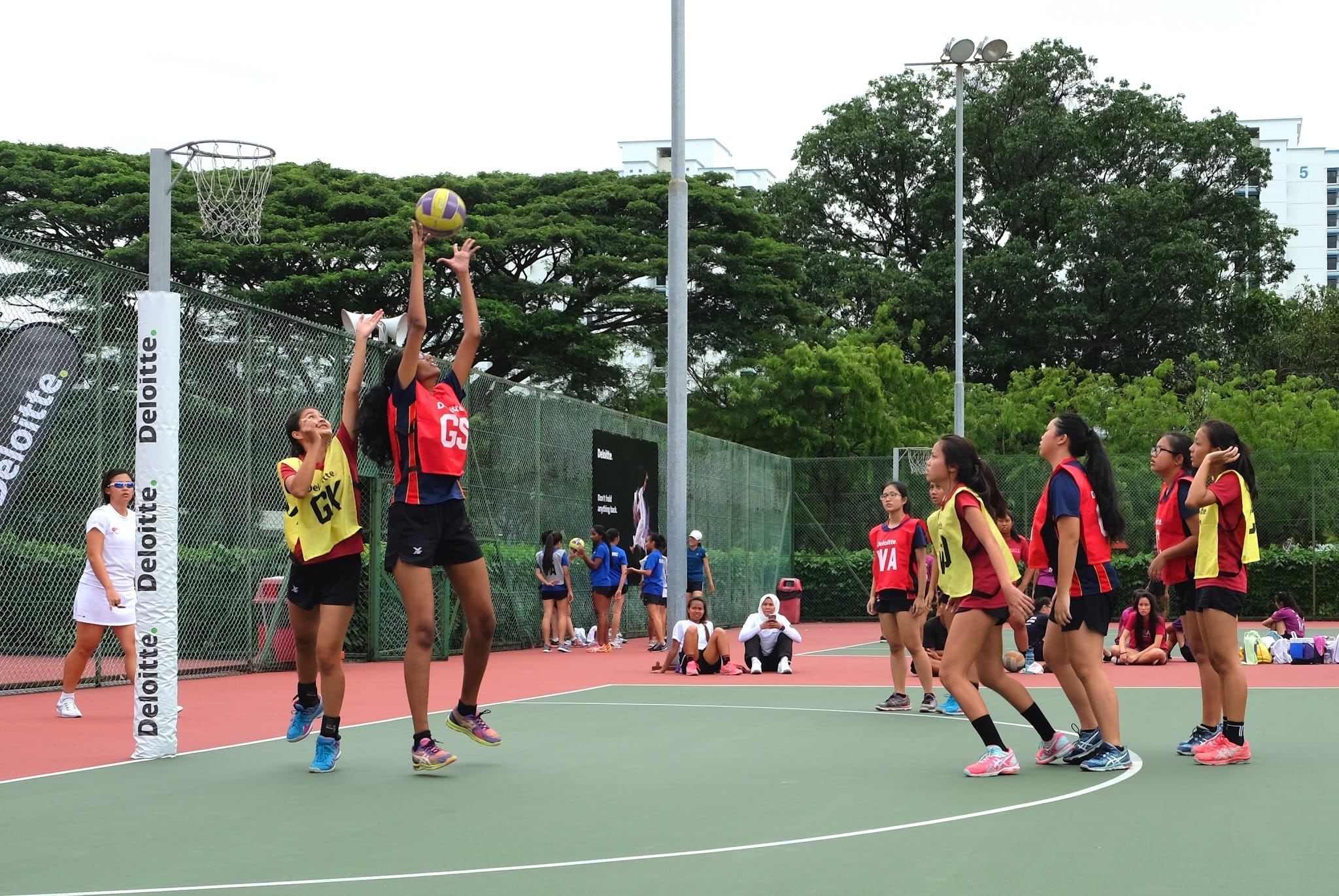 Girls playing netball outdoors, with one player jumping to shoot near the post while others defend and watch.