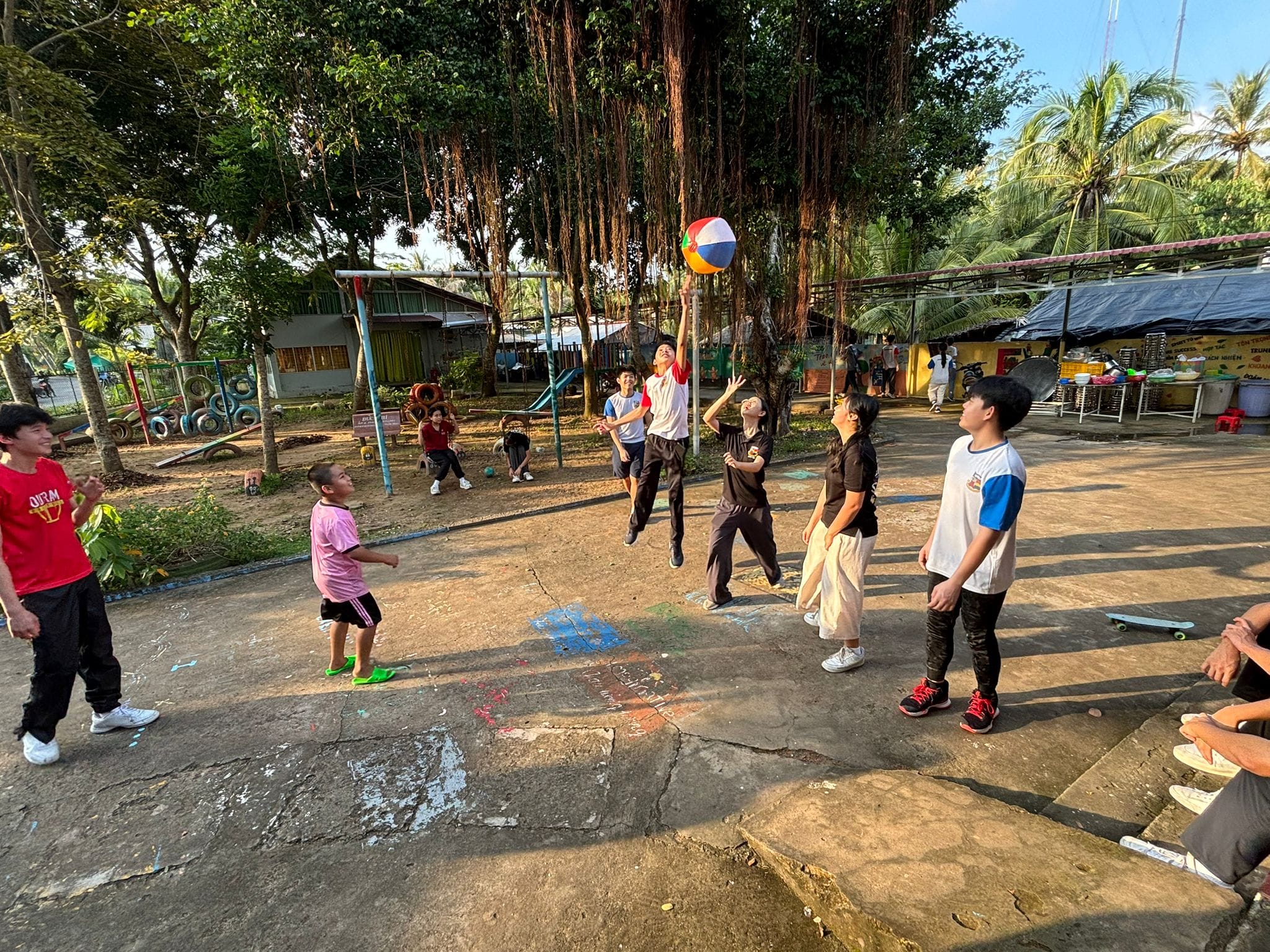 A group of children play with a colorful beach ball on an outdoor concrete court surrounded by large trees.