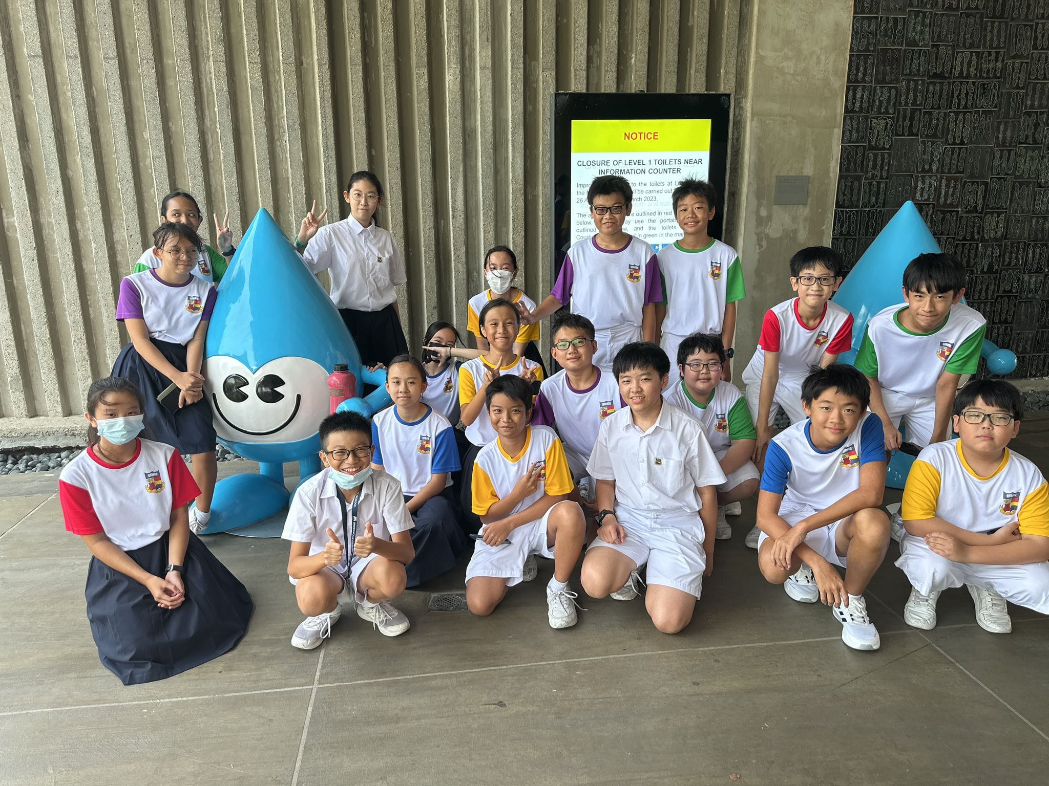 Group of school children in uniforms posing outdoors beside a blue smiling water drop mascot statue.