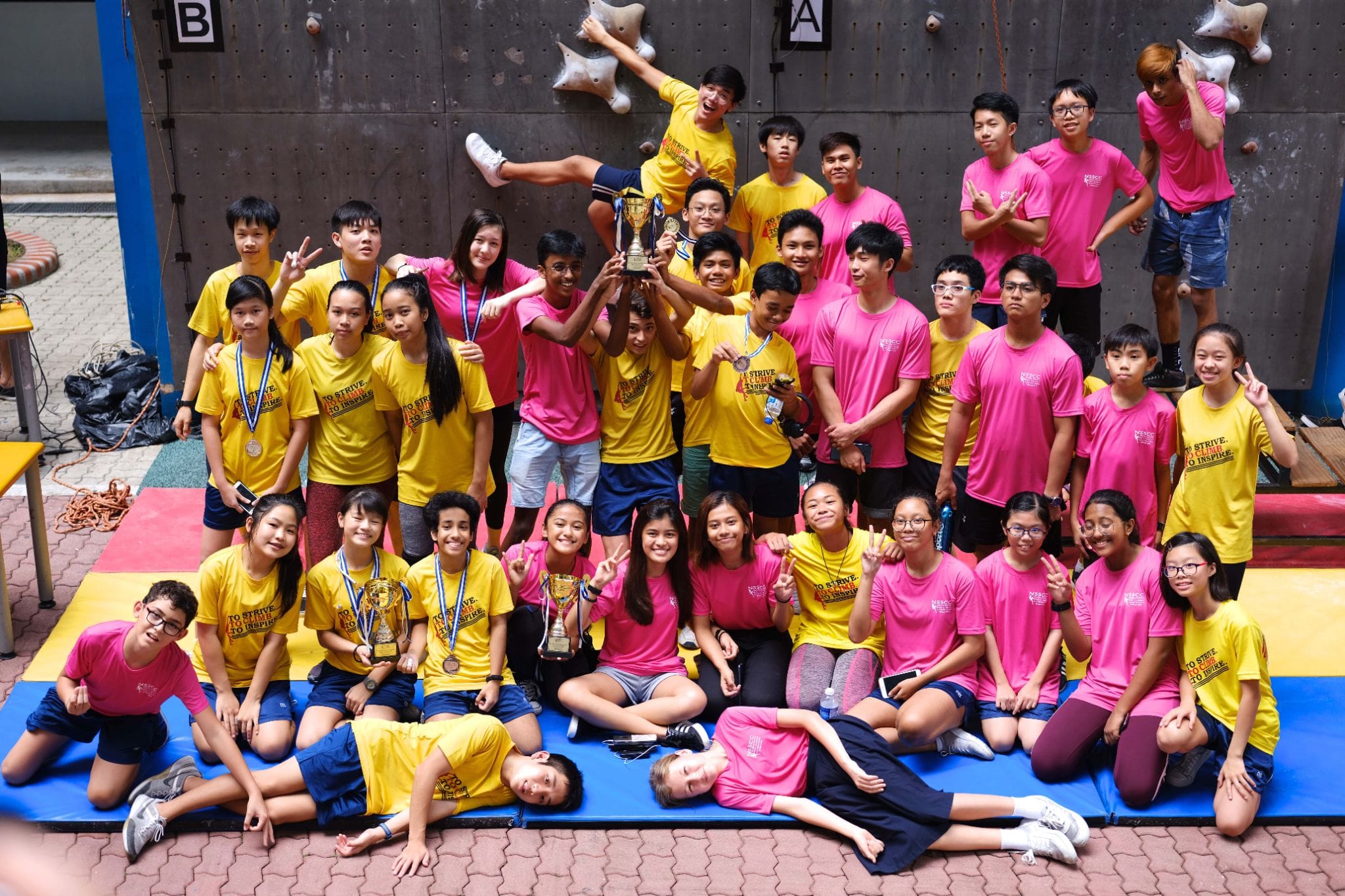 Large group of students in yellow and pink shirts posing with trophies and medals in front of a climbing wall.