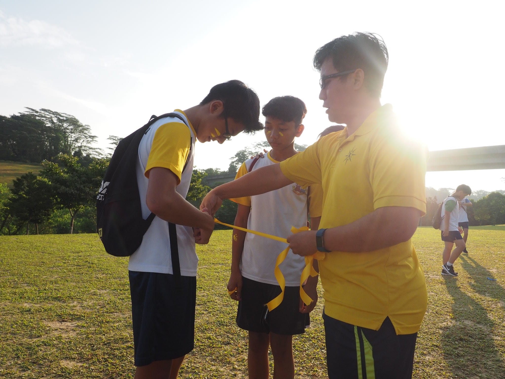An adult in a yellow shirt ties a yellow ribbon on a boy's wrist while another boy watches on a grassy field.