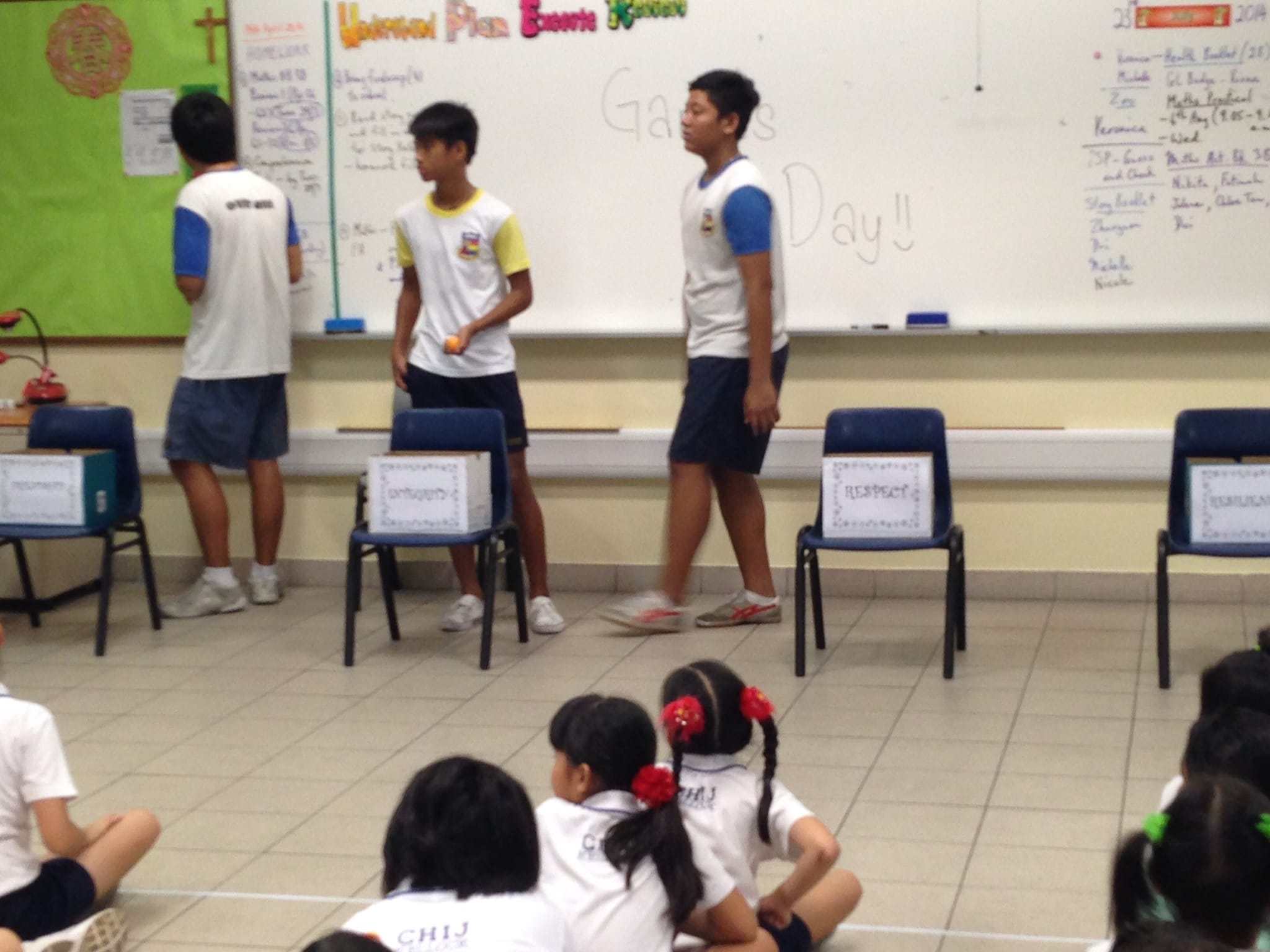 Three students in uniforms stand near a whiteboard in a classroom while younger students sit on the floor watching.
