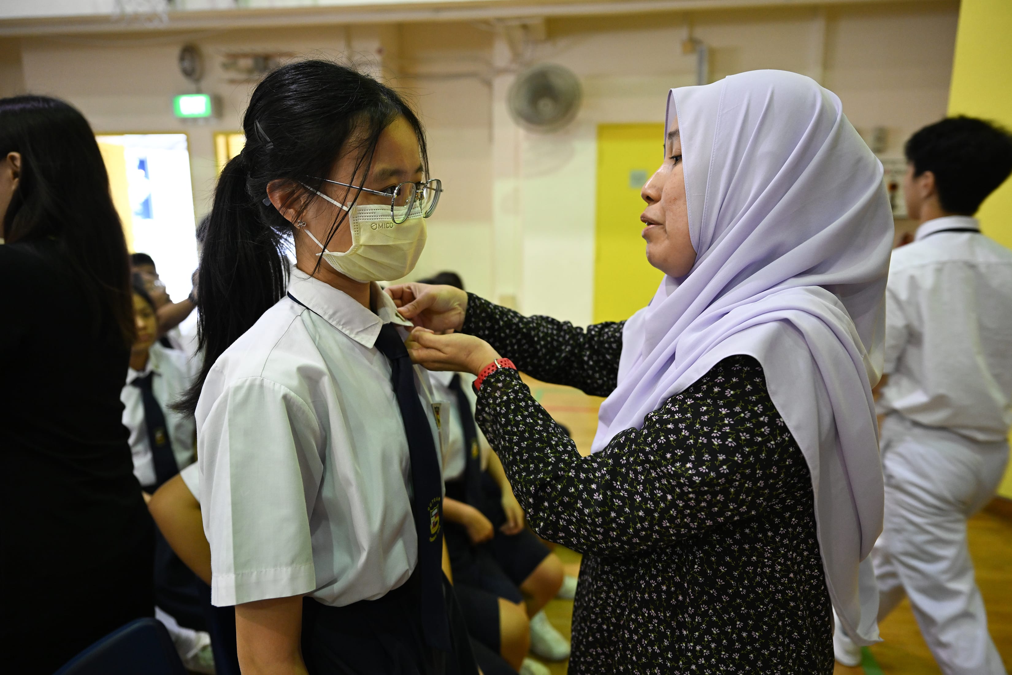 A lady in black and white dress helping a girl in white uniform wear a badge on her collar.