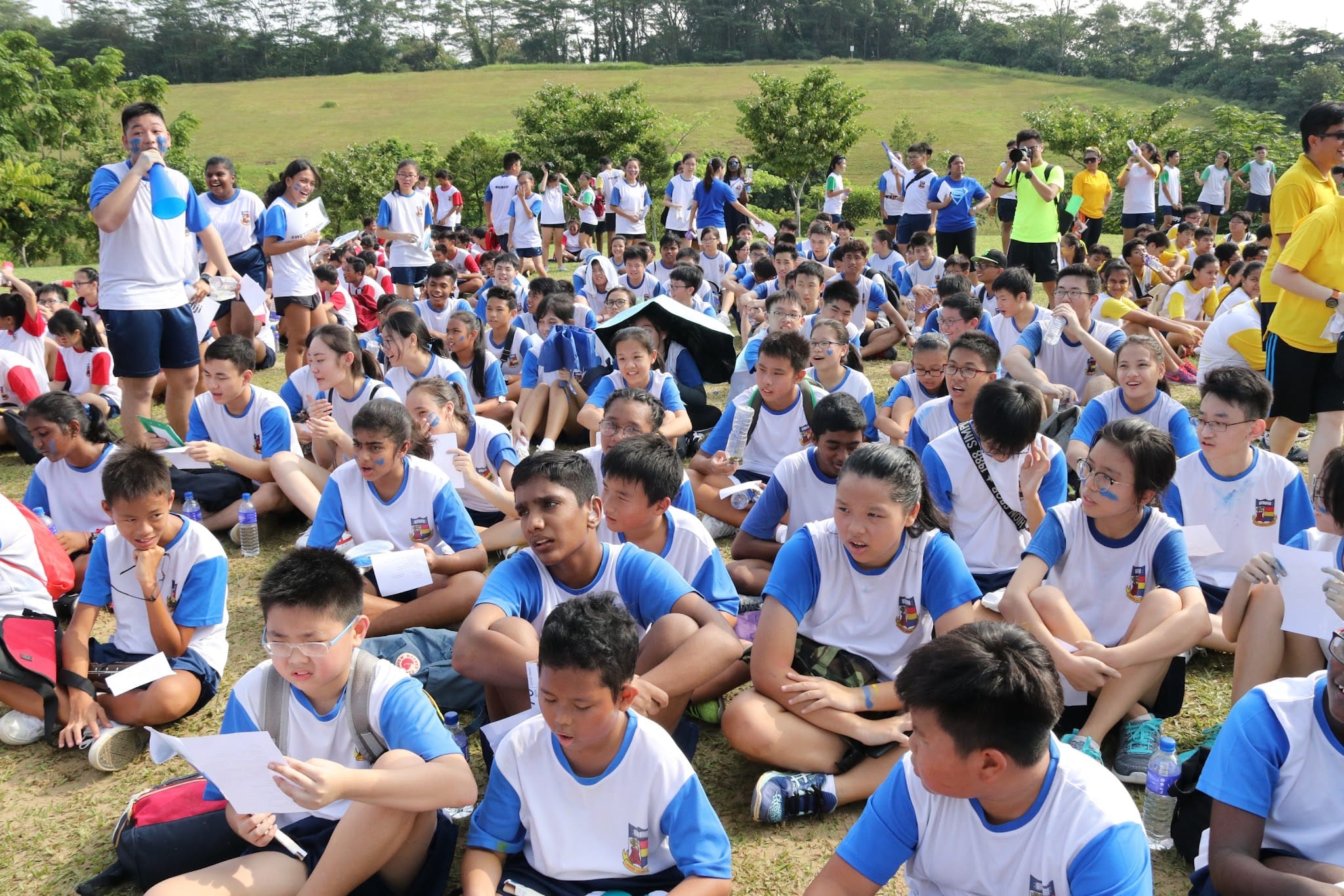 A large group of students in white and blue uniforms sit on grass outdoors during a school event.